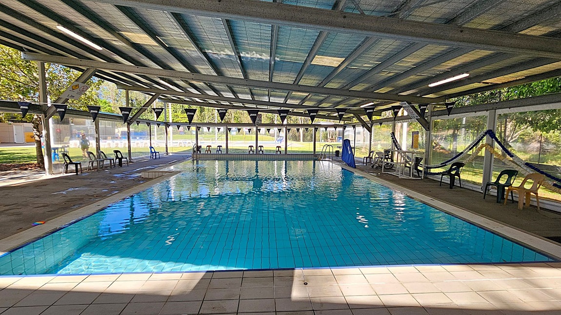 An indoor swimming pool with clear blue water, surrounded by a tiled floor. The ceiling is made of metal beams with translucent panels allowing natural light to filter through. On the left and right sides, there are plastic chairs placed along the pool's edge. The background features large windows with views of a grassy outdoor area and trees. Bunting with the word "Zoggs" is strung across the pool area. A lifeguard chair and some pool equipment are visible on the right side. An indoor swimming pool with clear blue water, surrounded by a tiled floor. The ceiling is made of metal beams with translucent panels allowing natural light to filter through. On the left and right sides, there are plastic chairs placed along the pool's edge. The background features large windows with views of a grassy outdoor area and trees. Bunting with the word "Zoggs" is strung across the pool area. A lifeguard chair and some pool equipment are visible on the right side.