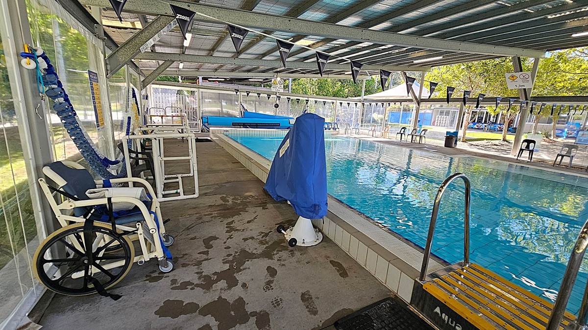 An indoor swimming pool area with a metal roof and transparent walls. On the left, there is a wheelchair and a blue pool lift. In the middle, the pool is visible with clear water, and a covered pool lift is positioned at the pool's edge. On the right, there are several plastic chairs lined up along the poolside. The floor is concrete with wet patches, and the ceiling has metal beams and lighting fixtures. Outside, trees and a playground area are visible through the transparent walls. An indoor swimming pool area with a metal roof and transparent walls. On the left, there is a wheelchair and a blue pool lift. In the middle, the pool is visible with clear water, and a covered pool lift is positioned at the pool's edge. On the right, there are several plastic chairs lined up along the poolside. The floor is concrete with wet patches, and the ceiling has metal beams and lighting fixtures. Outside, trees and a playground area are visible through the transparent walls.