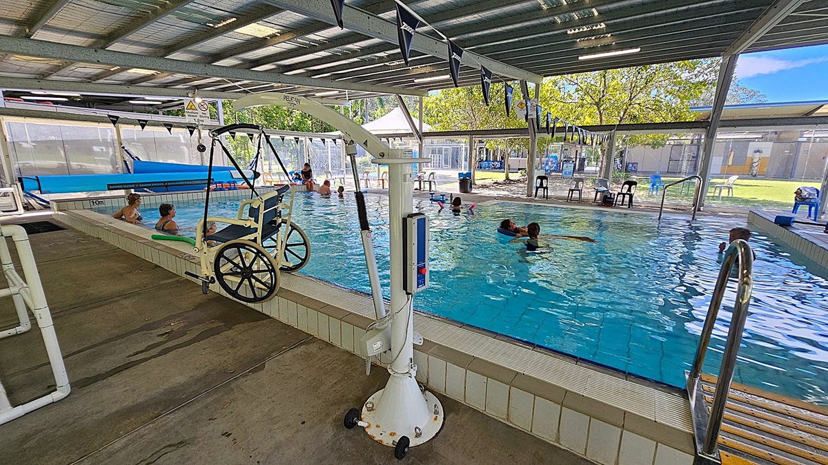 An indoor swimming pool with a metal roof and open sides. On the left, there is a pool lift with a wheelchair attached, and two people are sitting on the pool edge. In the middle, several people are swimming and engaging in water activities. On the right, there are metal handrails leading into the pool and a few plastic chairs and tables near the poolside. The floor is concrete, and the ceiling is made of metal sheets. Trees and a grassy area are visible outside. An indoor swimming pool with a metal roof and open sides. On the left, there is a pool lift with a wheelchair attached, and two people are sitting on the pool edge. In the middle, several people are swimming and engaging in water activities. On the right, there are metal handrails leading into the pool and a few plastic chairs and tables near the poolside. The floor is concrete, and the ceiling is made of metal sheets. Trees and a grassy area are visible outside.