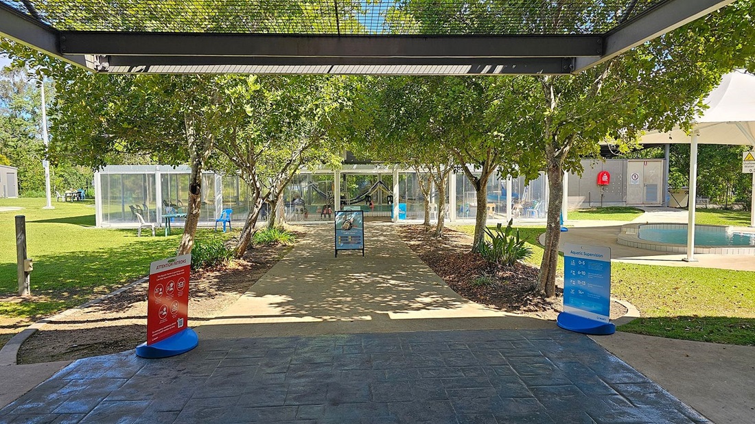 A paved pathway leads through a garden area with trees on both sides. In the foreground, there are two signs, one red and one blue, providing information. On the left, there is a grassy area with a transparent structure containing chairs. On the right, there is a small pool with a white canopy overhead. In the background, a glass building with people inside is visible. The floor is paved with stone tiles, and the ceiling is a metal grid structure. A paved pathway leads through a garden area with trees on both sides. In the foreground, there are two signs, one red and one blue, providing information. On the left, there is a grassy area with a transparent structure containing chairs. On the right, there is a small pool with a white canopy overhead. In the background, a glass building with people inside is visible. The floor is paved with stone tiles, and the ceiling is a metal grid structure.