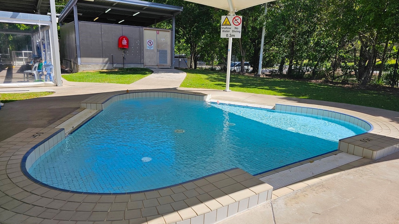 A small, circular wading pool is in the centre, covered by a white canopy supported by four poles. Surrounding the pool is a concrete pathway, with grass and trees on either side. To the left, there is a glass-walled building with chairs inside and a red fire extinguisher mounted on the exterior wall. In the foreground, a blue sign with "Aquatic Supervision" guidelines is visible. To the right, there are a few plastic chairs and a fence with parked cars behind it. A small, circular wading pool is in the centre, covered by a white canopy supported by four poles. Surrounding the pool is a concrete pathway, with grass and trees on either side. To the left, there is a glass-walled building with chairs inside and a red fire extinguisher mounted on the exterior wall. In the foreground, a blue sign with "Aquatic Supervision" guidelines is visible. To the right, there are a few plastic chairs and a fence with parked cars behind it.
