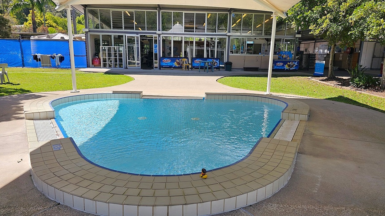 A small, shallow swimming pool with a tiled edge and a rubber duck floating in the water. The pool is surrounded by a concrete walkway. In the background, there is a building with large glass windows and doors, displaying signs for "Peters" ice cream. To the left, there is a grassy area with a white plastic chair and clothes hanging on a line. To the right, there are trees and more grassy areas. A small, shallow swimming pool with a tiled edge and a rubber duck floating in the water. The pool is surrounded by a concrete walkway. In the background, there is a building with large glass windows and doors, displaying signs for "Peters" ice cream. To the left, there is a grassy area with a white plastic chair and clothes hanging on a line. To the right, there are trees and more grassy areas.