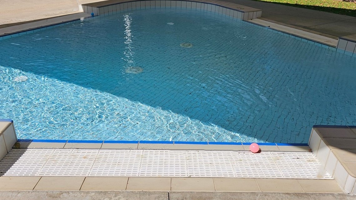 A swimming pool with clear blue water. The pool has a tiled edge and a shallow end marked "0.3m". On the poolside, there are a few leaves and a pink ball. The surrounding area includes a concrete walkway and a grassy area in the background. The pool is partially shaded by a structure. A swimming pool with clear blue water. The pool has a tiled edge and a shallow end marked "0.3m". On the poolside, there are a few leaves and a pink ball. The surrounding area includes a concrete walkway and a grassy area in the background. The pool is partially shaded by a structure.