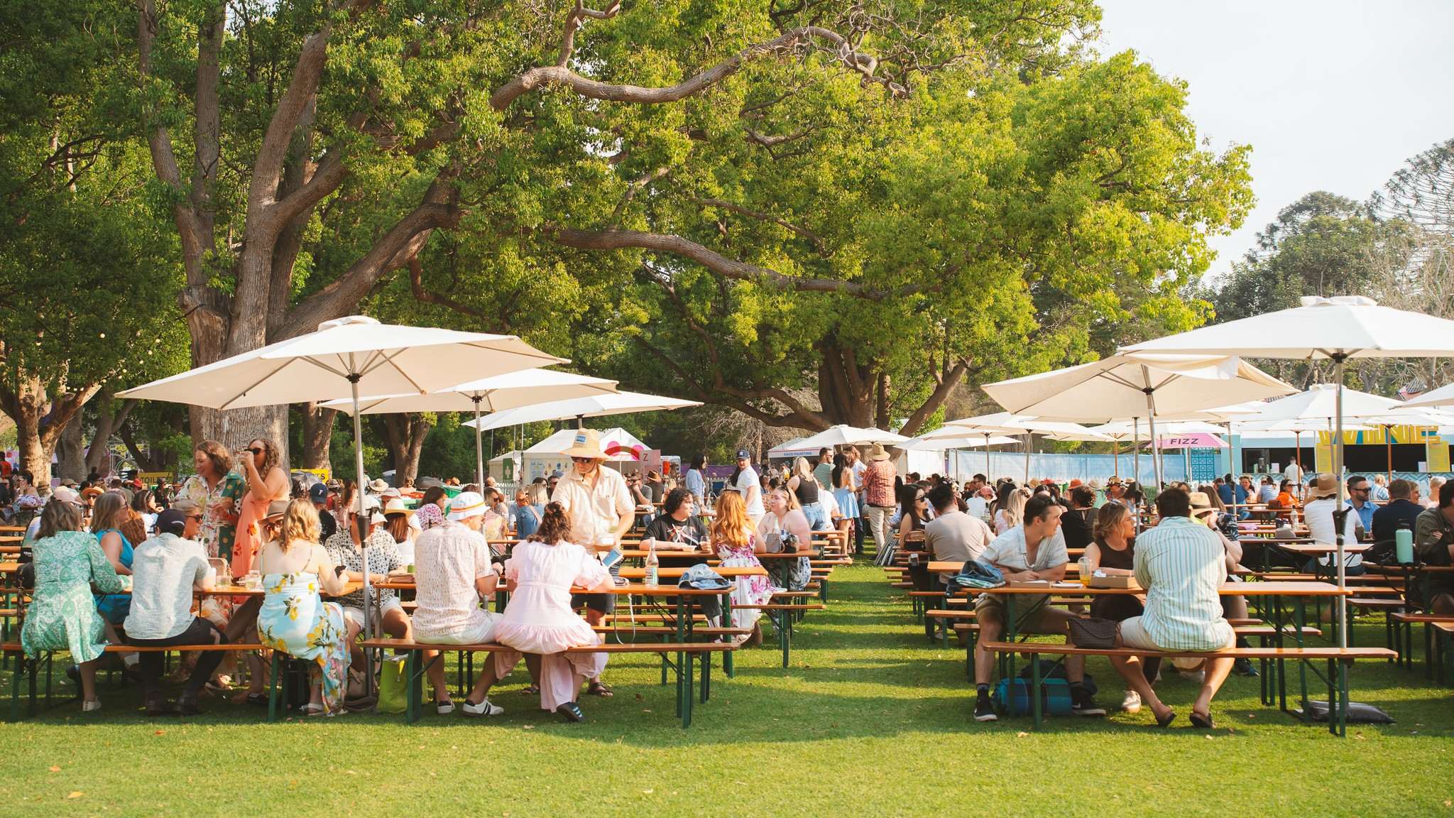 People sit at outdoor picnic tables under large umbrellas at a busy event, with food stalls, marquees and mature trees surrounding the area.
