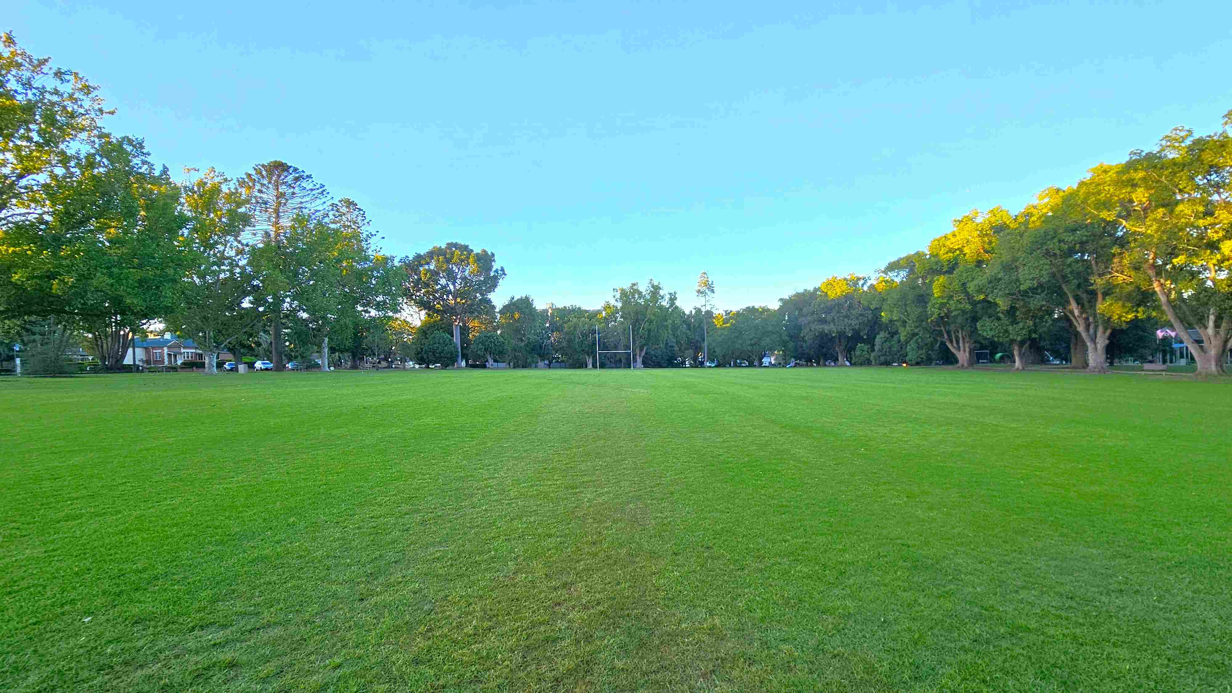 Large open grassed field surrounded by mature trees, with a broad, mostly flat lawn under a clear blue sky.