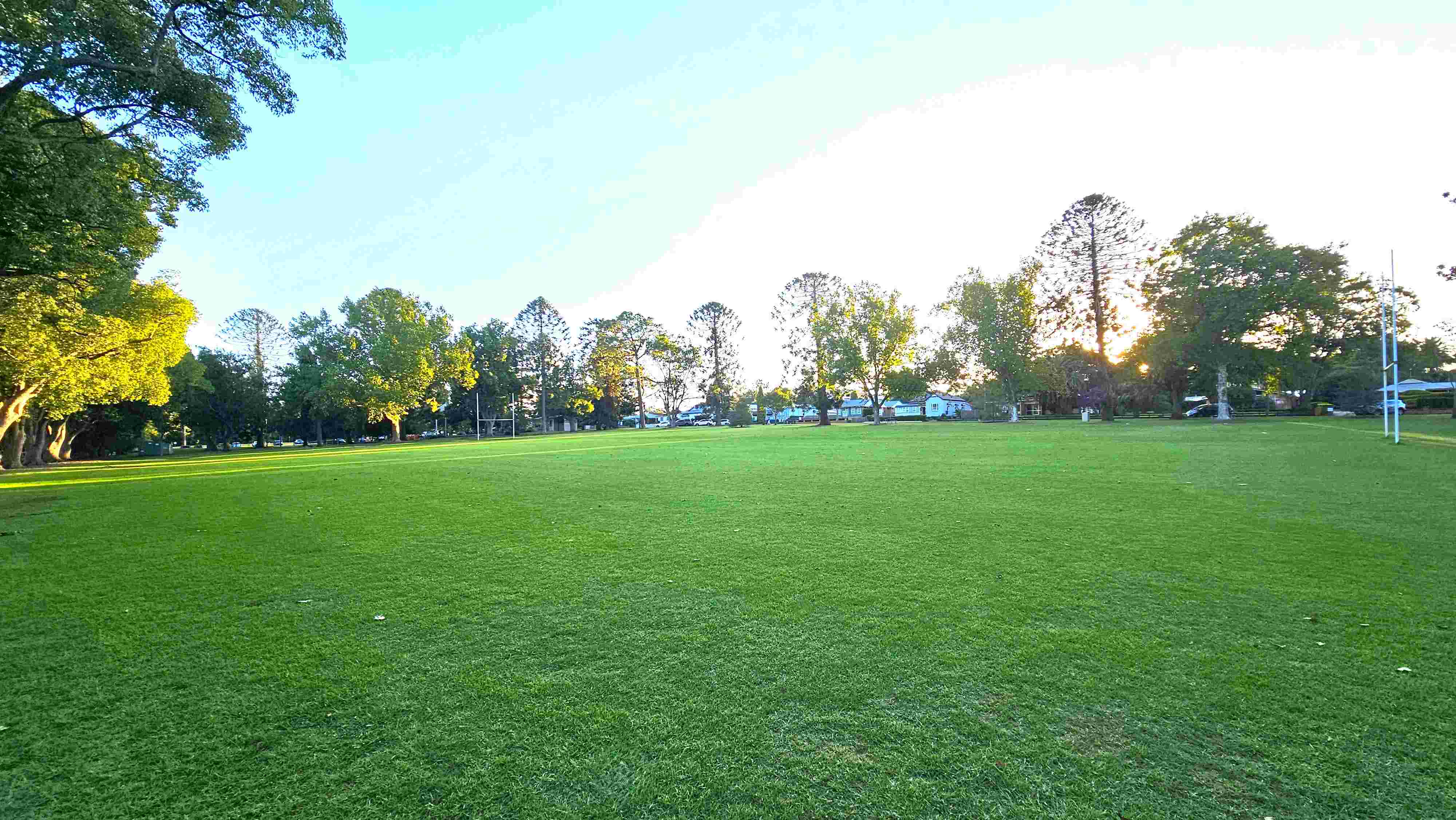 Wide open grassed field bordered by mature trees, with a mostly flat lawn and buildings visible through the trees in the distance.