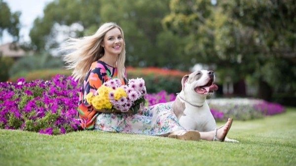 Woman and small dog sit together on a grassed lawn beside a garden bed filled with bright pink flowers.