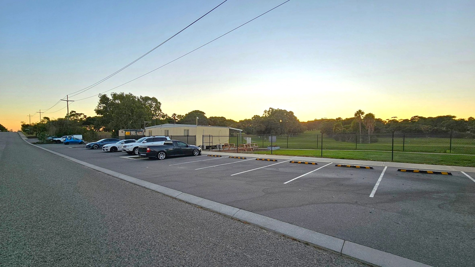 A paved road runs along the left side, with a row of parked cars adjacent to it. On the right, there is an empty parking area with marked spaces. In the middle, a small building with a light-coloured exterior is visible, surrounded by a fence. Behind the building, there is a grassy field with scattered trees. The sky is transitioning from day to night, with a gradient of colours near the horizon.