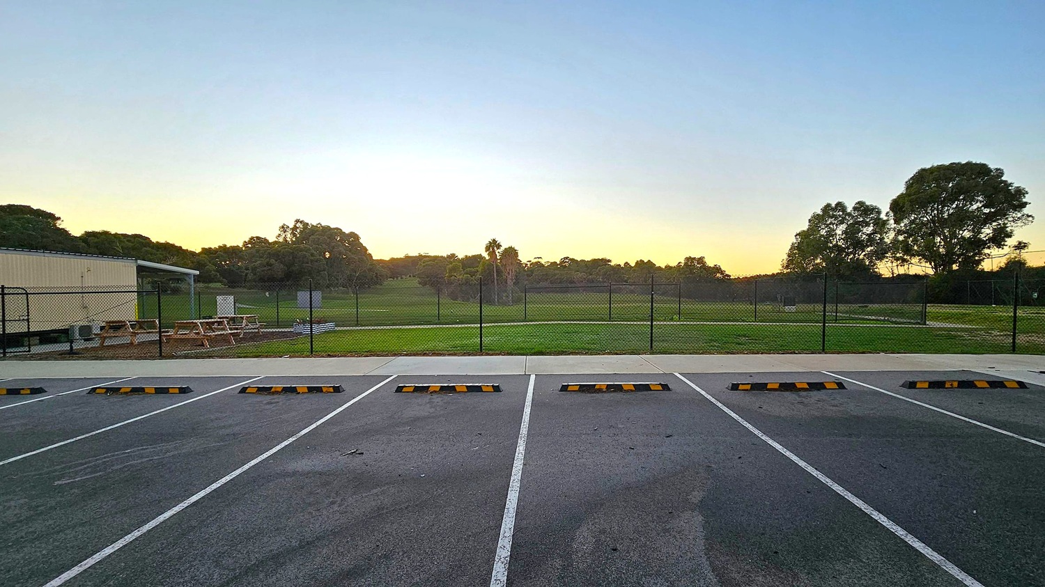 A row of empty parking spaces with yellow and black wheel stops in the foreground. Beyond the parking area, a chain-link fence encloses a grassy field. To the left, there are picnic tables and a small structure. Trees are scattered across the background, with a few taller ones silhouetted against the sky. The ground is paved, and the sky has a gradient from light to darker blue.