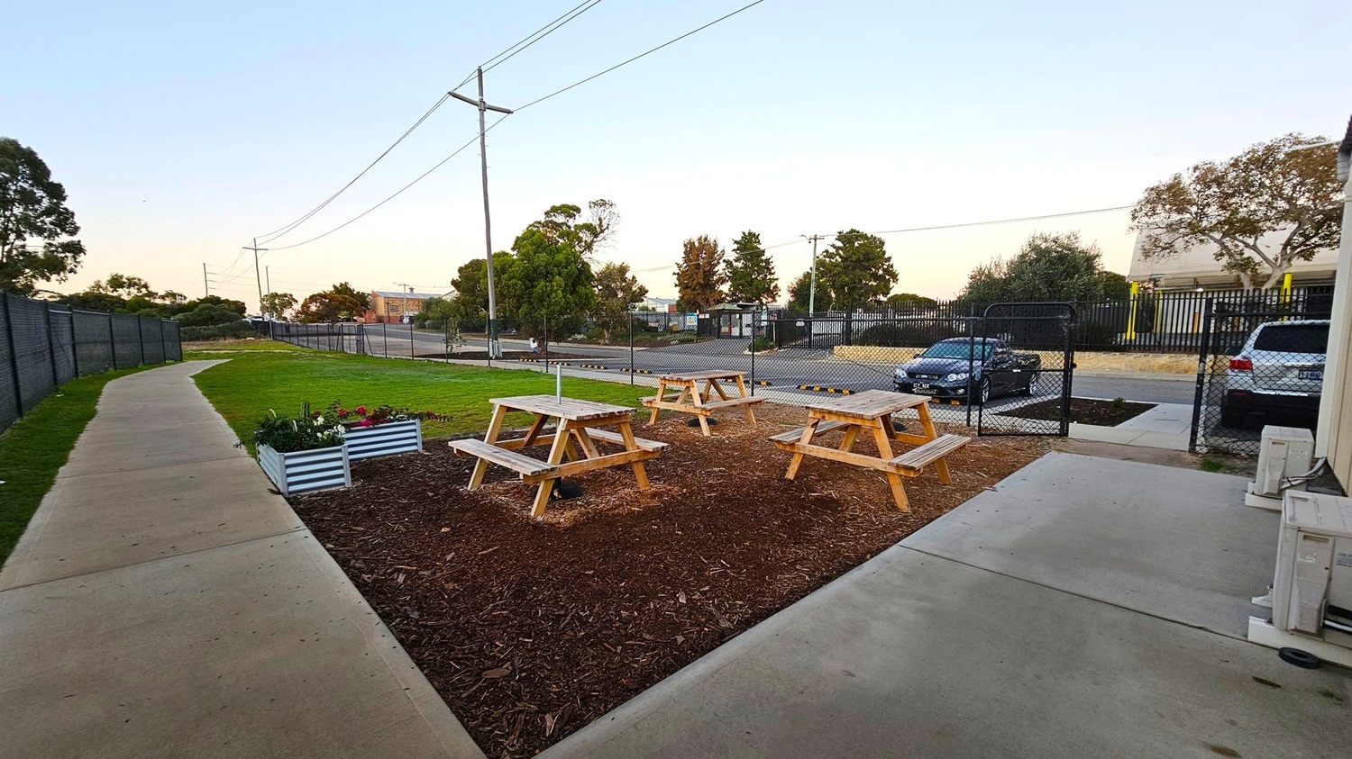 A small outdoor area with three wooden picnic tables on a patch of mulch. To the left, a concrete path runs alongside a grassy area with a few raised flower beds. In the background, a chain-link fence separates the area from a road where two cars are parked. Trees and utility poles line the street, and a building is visible in the distance. The ground is a mix of concrete, grass, and mulch.