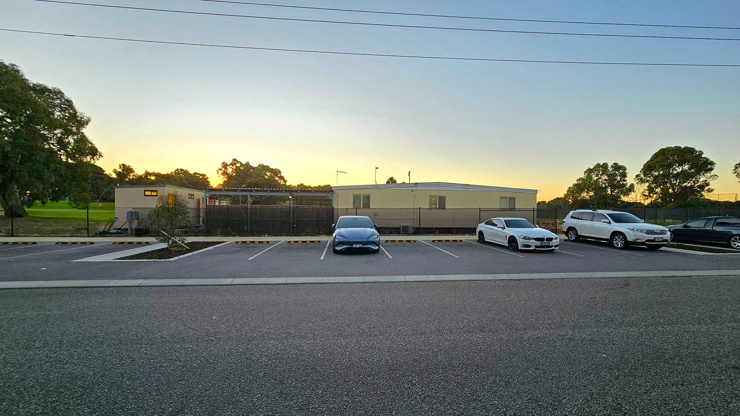 A parking area with several cars lined up. In the centre, a blue car is parked, flanked by a white car on the right and another white vehicle further to the right. On the far right, a dark-coloured car is visible. In the background, a single-storey building with a flat roof and small windows is surrounded by a fence. Trees are visible on both sides, with a grassy area on the left. The ground is paved, and there are parking space markers.