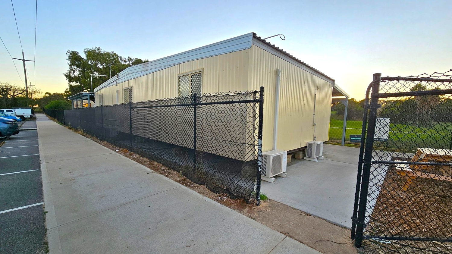 A long, light-coloured portable building with a corrugated metal roof is on the right, surrounded by a black chain-link fence. Two air conditioning units are visible at the base. A concrete path runs alongside the building, leading to a parking area on the left with several parked cars. In the background, there are trees and a grassy field. The sky is clear, and the area is well-lit, suggesting early morning or late afternoon.
