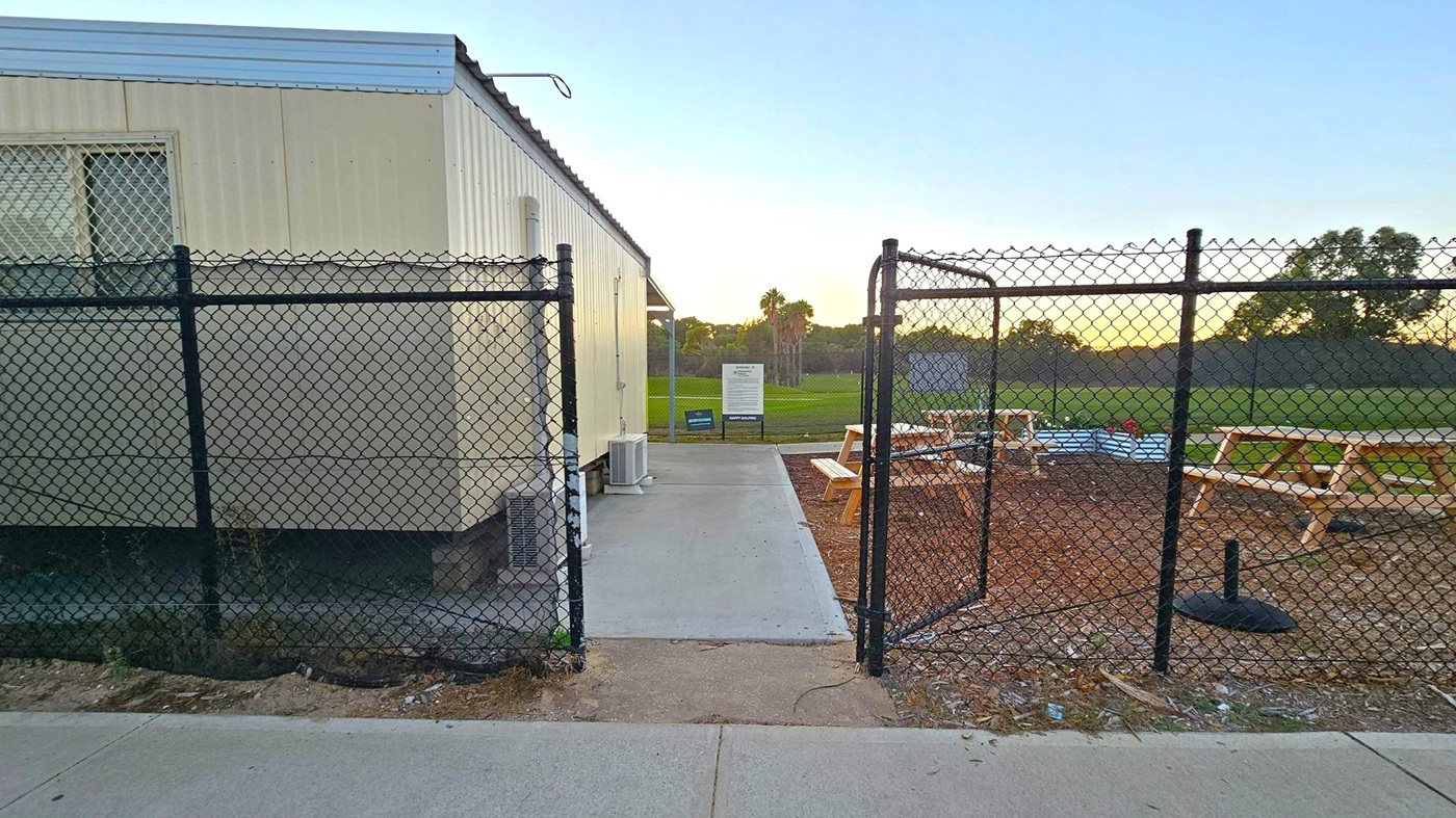 A chain-link fence with an open gate is in the foreground, leading to a concrete path. On the left, a light-coloured portable building with a barred window and an air conditioning unit is visible. To the right, there are wooden picnic tables on a dirt area. In the background, a grassy field with trees and a sign can be seen. The sky is light, suggesting early morning or late afternoon. The ground is a mix of concrete and dirt.