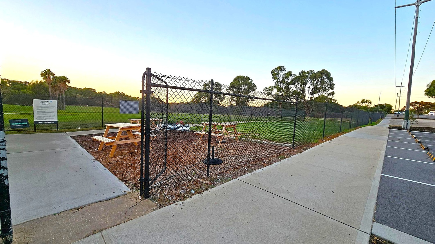 A fenced area with picnic tables on the left, surrounded by grass and trees in the background. A sign is visible near the entrance. On the right, a concrete footpath runs alongside a parking area with marked spaces. Tall utility poles line the path, and the sky is clear.