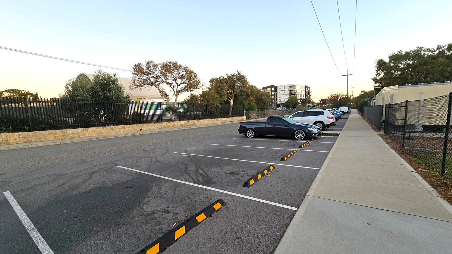 A parking area with several cars parked on the right side, including a black vehicle in the foreground. The left side features a stone wall and a black metal fence with trees and shrubs behind it. In the background, a multi-storey building is visible. A footpath runs parallel to the parking spaces on the right, bordered by a black chain-link fence. Overhead, power lines stretch across the scene. The ground is asphalt, and the footpath is concrete with a green strip.