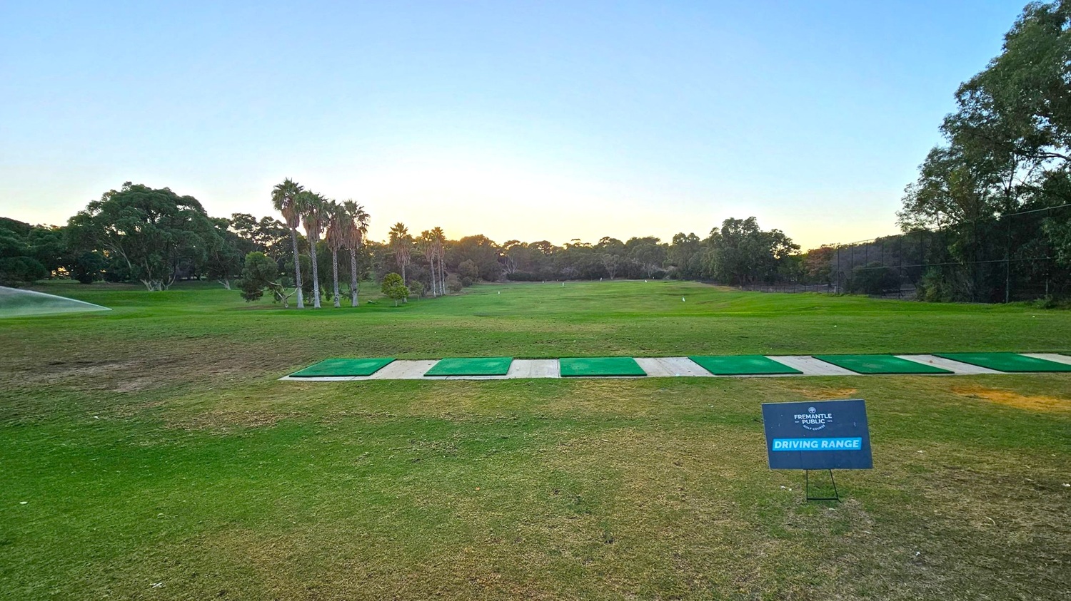 A golf driving range with a grassy field extending into the distance. In the foreground, there are several green mats lined up for practice. A sign on the right reads "Fremantle Public Golf Course Driving Range." On the left, a sprinkler is watering the grass. Tall trees, including palm trees, are scattered across the background, with a clear sky above.