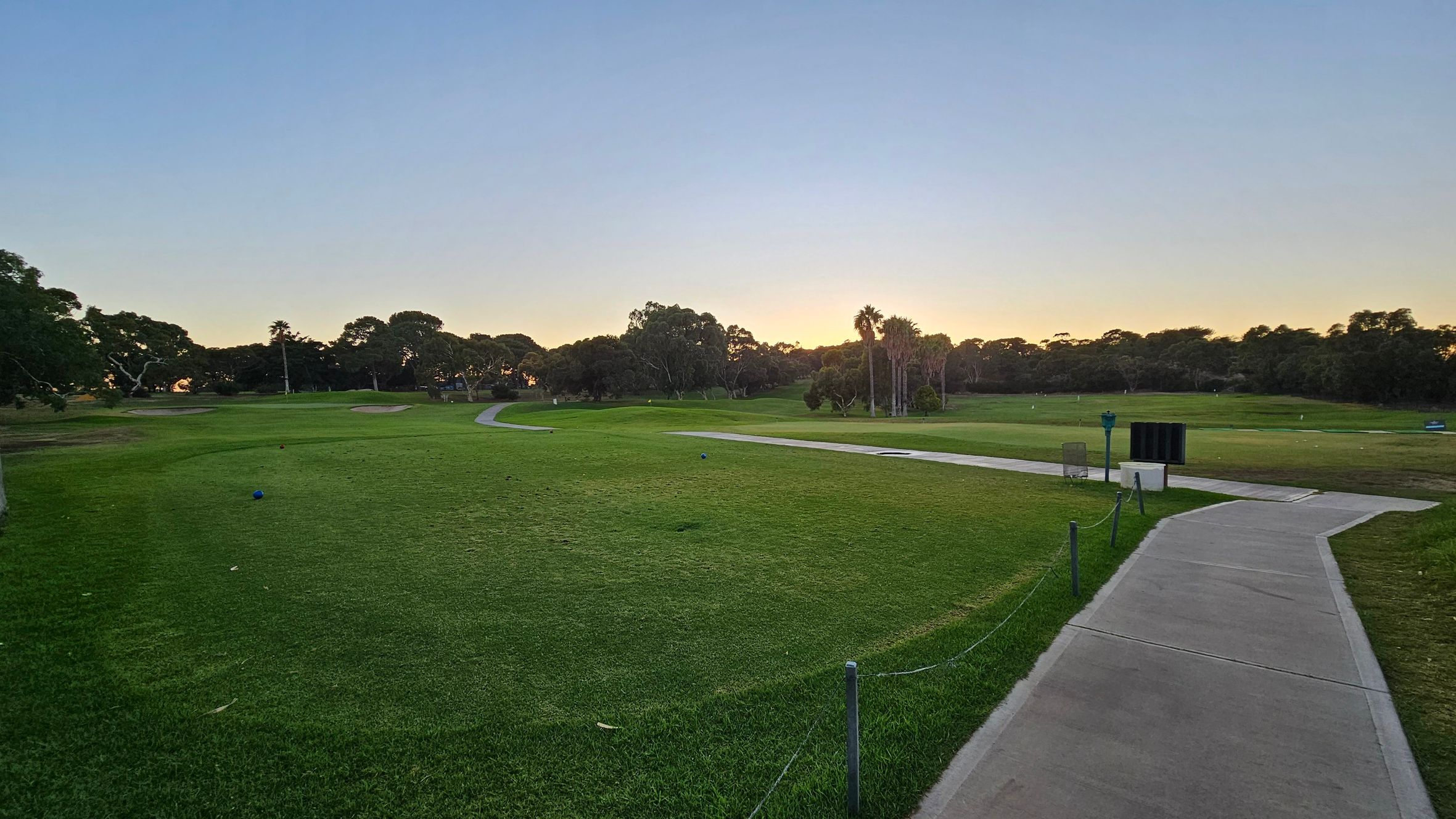 A golf course with a well-maintained green landscape. A concrete path runs from the foreground to the right, bordered by a low rope fence. In the middle, there are scattered trees and a few palm trees. The horizon shows a line of trees silhouetted against a setting or rising sun, casting a warm glow. The sky is clear with a gradient from light to darker blue.