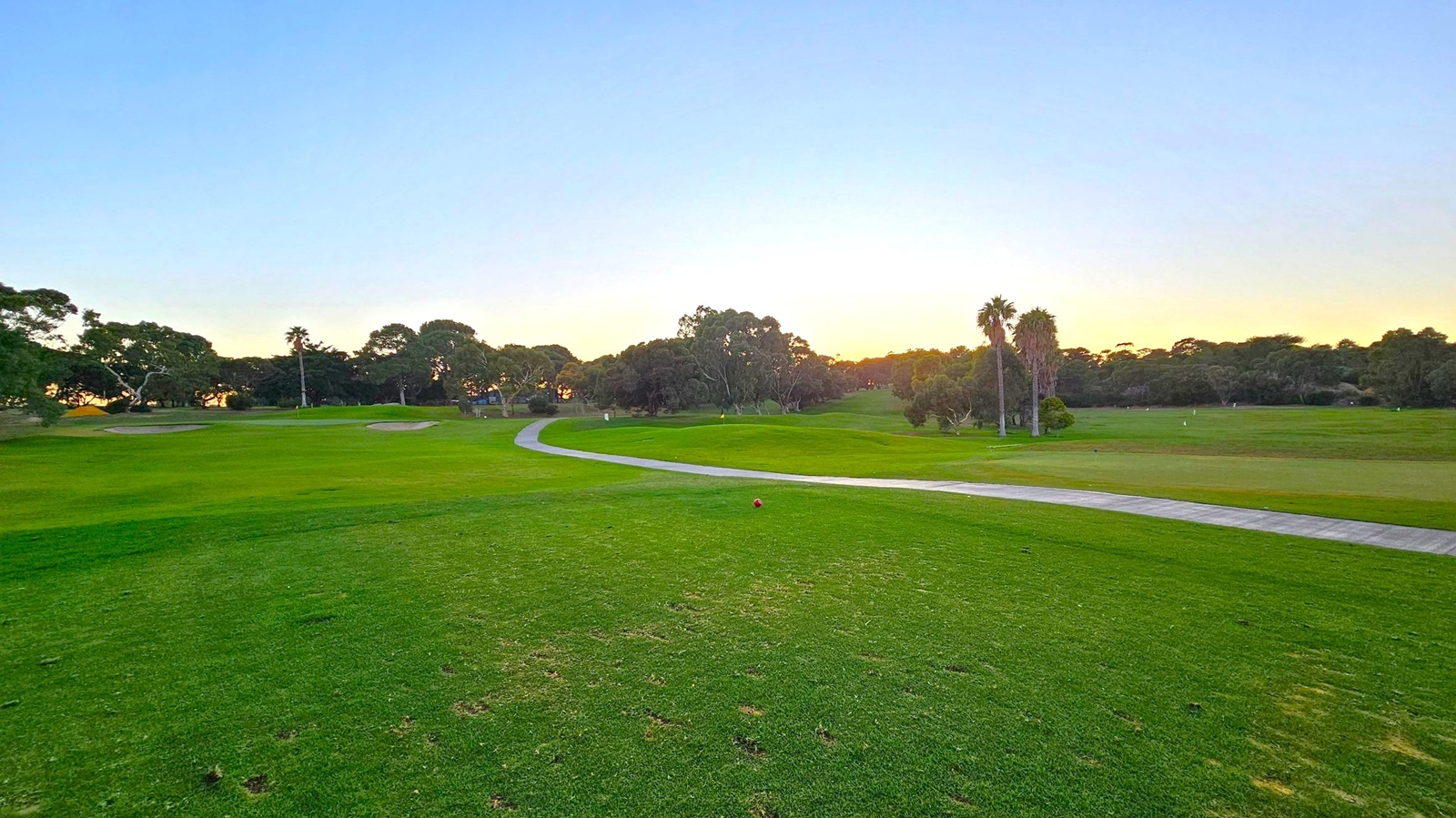 A golf course with a well-maintained green field stretches across the image. In the middle, a winding path leads through the course, flanked by trees and a few palm trees on the right. The foreground shows a patchy grass area with a red marker. The background is filled with dense trees, and the sun is setting, casting a warm glow over the landscape.