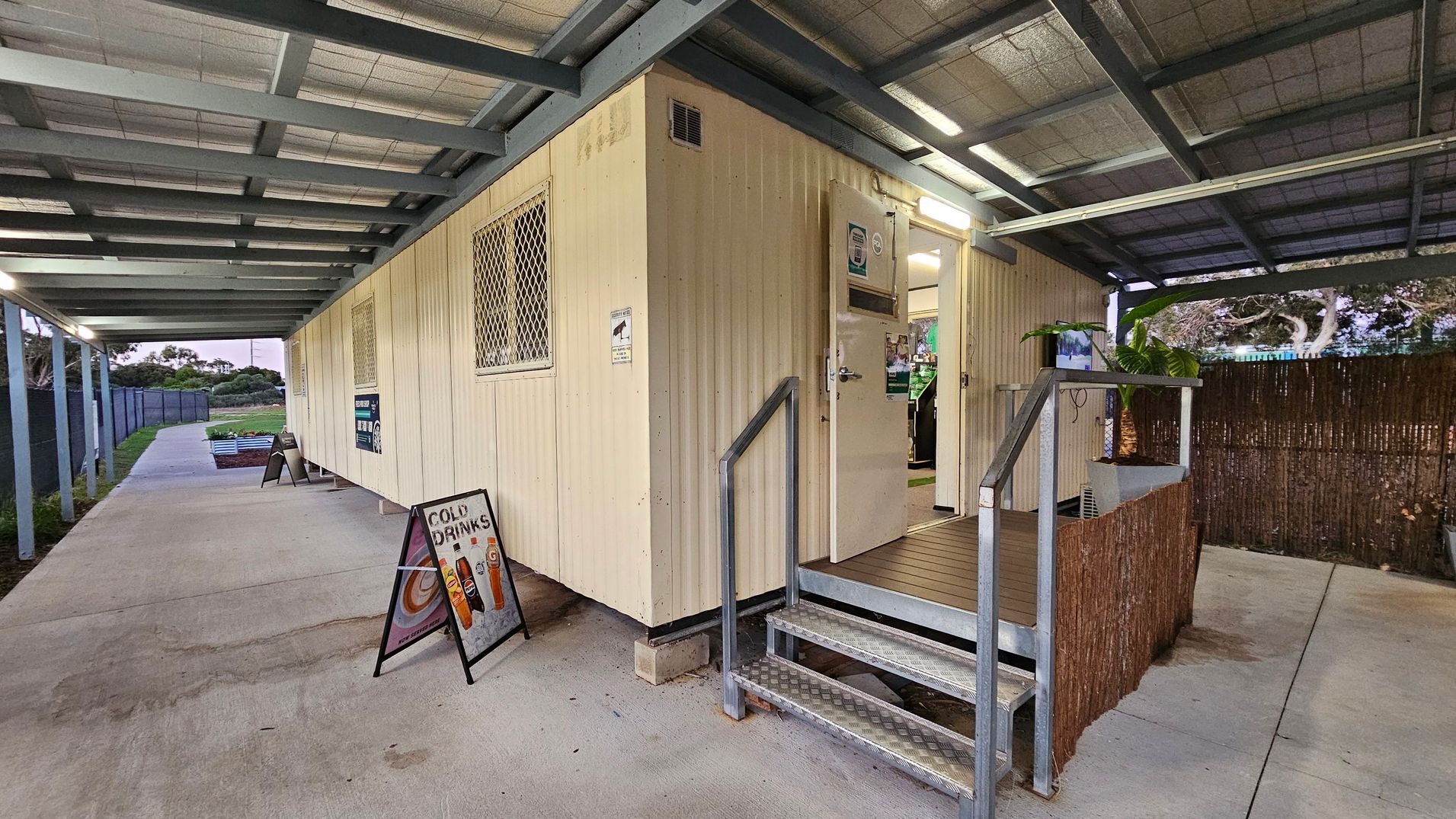 A small, beige portable building with a metal roof and steps leading to an open door. On the left, a concrete path runs alongside the building, with a sign advertising cold drinks. The building has barred windows and a few signs on its exterior. To the right, a wooden fence and a potted plant are visible. The ceiling is metal with exposed beams, and the floor is concrete. In the background, there are trees and a grassy area.