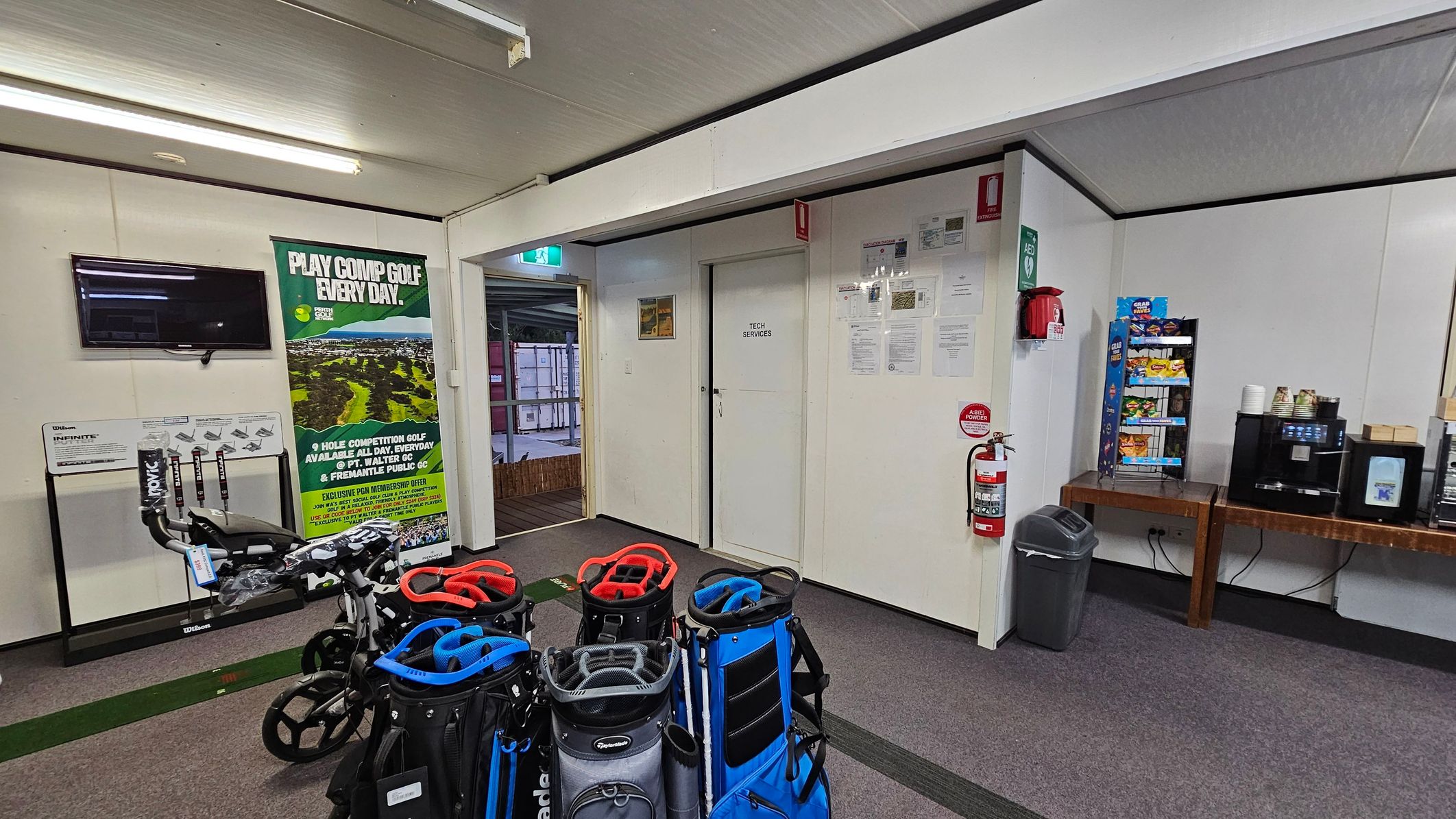 Golf bags with trolleys are in the foreground on a carpeted floor. On the left, a wall-mounted TV and a poster about competitive golf. In the middle, a door labelled "Tech Services" with notices and a fire extinguisher nearby. On the right, a snack display and a coffee machine on a wooden table. The ceiling is white with fluorescent lights.
