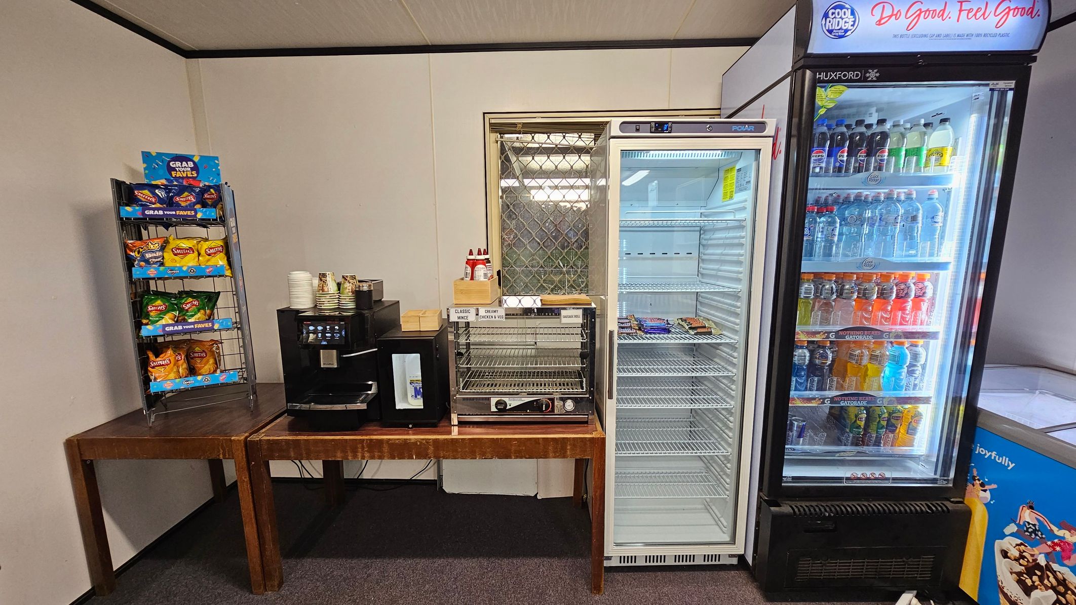 A small snack area with a variety of items. On the left, a metal rack holds packets of crisps. Next to it, a wooden table supports a coffee machine with stacked paper cups. In the centre, a heated display case contains a few food items. To the right, an empty glass-fronted fridge stands beside a stocked beverage cooler filled with bottled drinks. The floor is carpeted, and the ceiling is plain with visible panels.