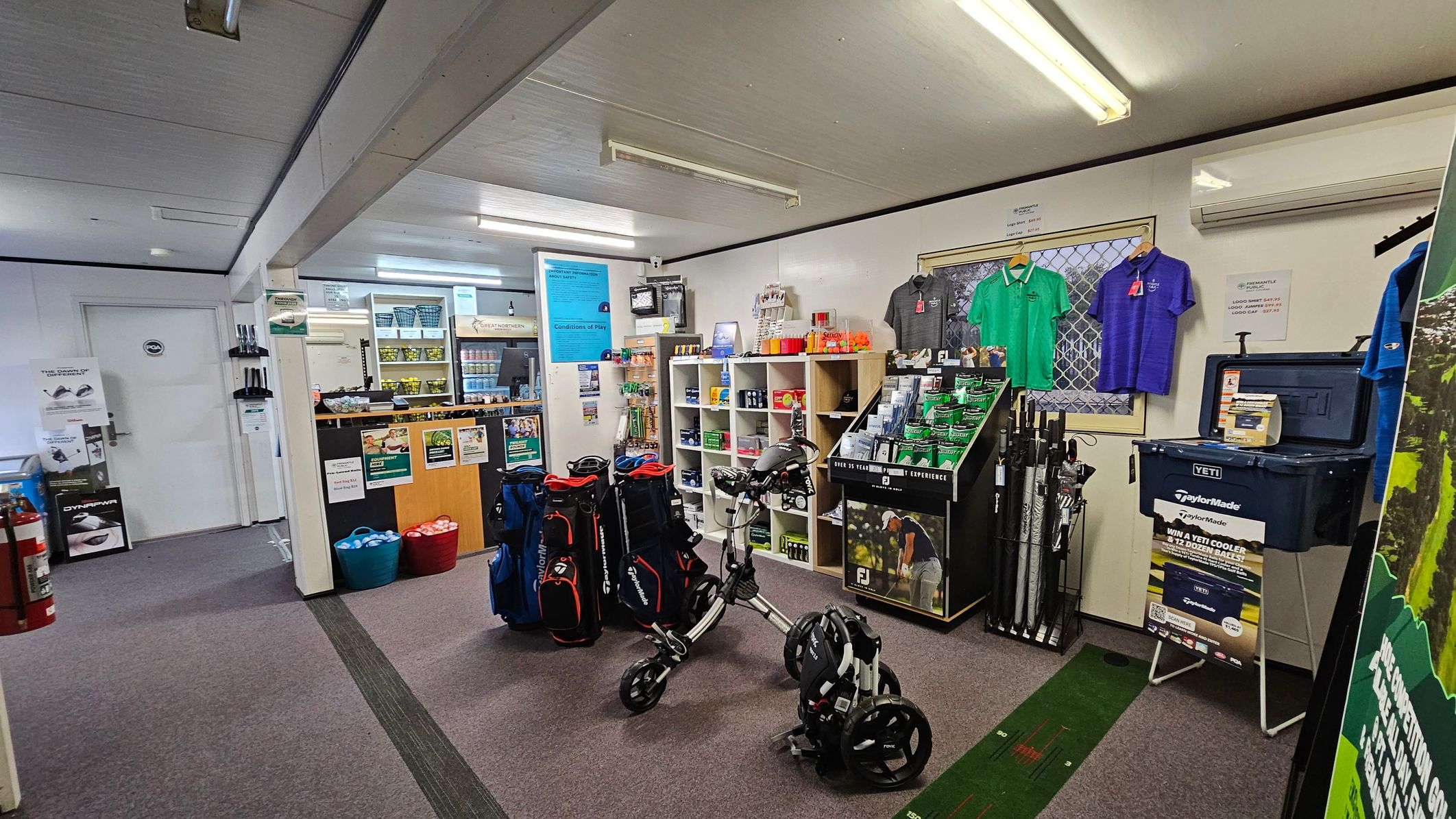 Golf shop interior with a grey carpeted floor and white ceiling. On the right, golf shirts hang on the wall above a display of golf accessories and a Yeti cooler. In the centre, a golf trolley and bags are displayed. The left side features a counter with various golf products, a door, and a fire extinguisher. Shelves behind the counter hold more items, and posters are visible on the walls. Fluorescent lights illuminate the space.