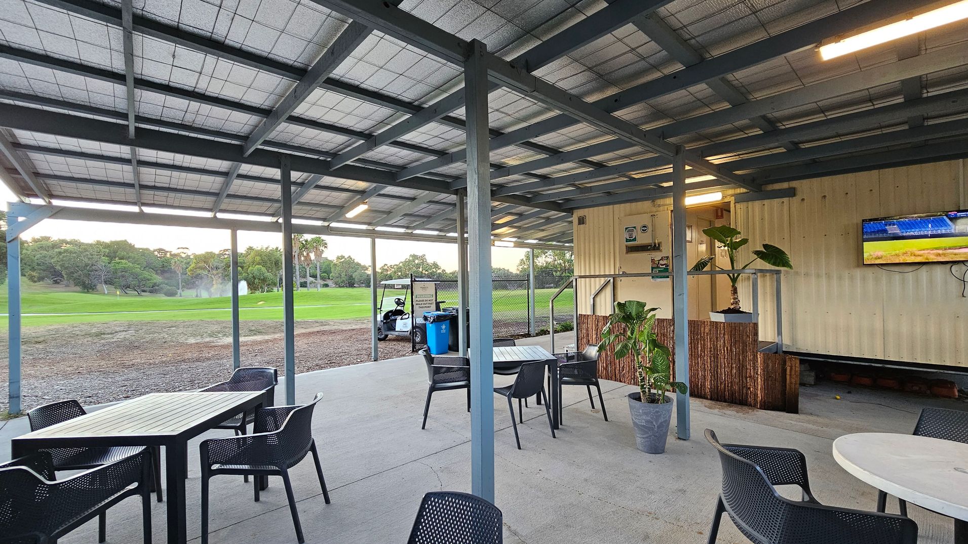 A covered outdoor seating area with a metal roof and concrete floor. On the left, there are tables and chairs arranged for dining. In the middle, a golf cart and bins are visible near a fence. On the right, a wall with a mounted television and potted plants. The background features a green golf course with trees and a water feature.