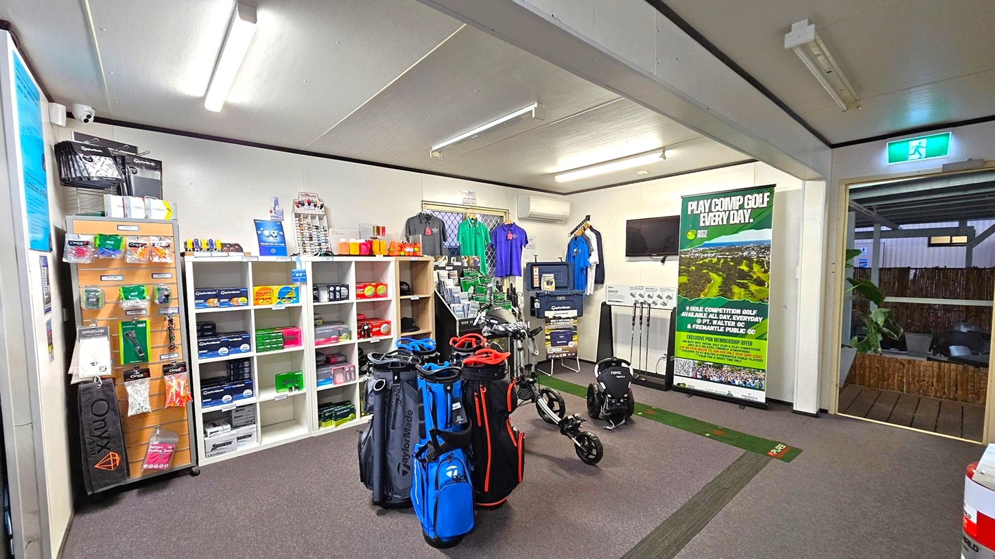 A golf shop interior with shelves on the left displaying various golf accessories like balls, tees, and gloves. In the centre, several golf bags in blue, black, and red are on display alongside a golf trolley. The back wall features a selection of polo shirts in different colours. A large poster promoting golf competitions is on the right, near a small TV mounted on the wall. The floor is carpeted, and the ceiling has fluorescent lighting. An exit sign is visible above a doorway on the right.