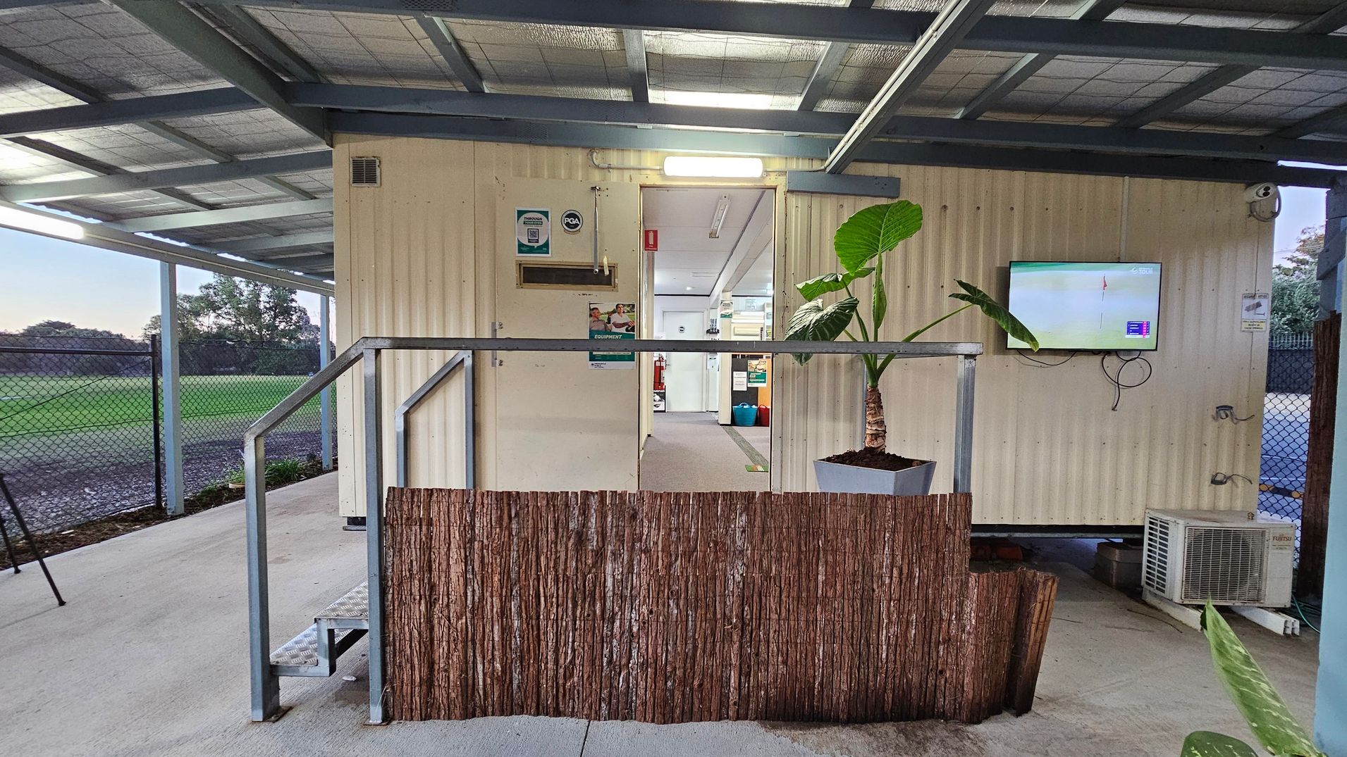 A small building with a corrugated metal exterior. In the centre, a doorway leads inside, revealing a corridor with fluorescent lighting. To the right, a mounted television screen displays an image, and a potted plant sits nearby. An air conditioning unit is positioned below. On the left, a metal railing and steps lead up to the entrance. The ceiling is metal, and the floor is concrete. A chain-link fence and grassy area are visible in the background.