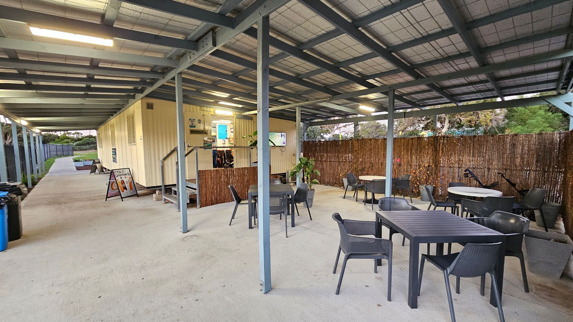 A covered outdoor seating area with a concrete floor and a metal roof. On the right, there are several tables with black chairs. In the middle, a small building with a service window and a sign advertising cold drinks. A plant is placed near the window. On the left, a walkway leads to a grassy area, with bins placed along the path. The background features a fence and some trees.