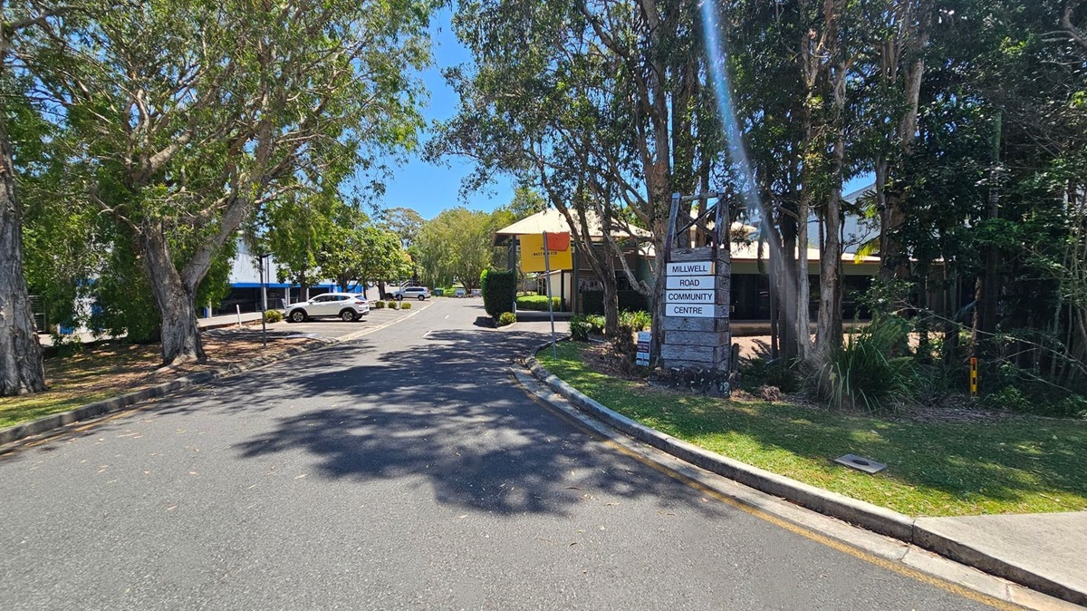 A tree-lined road with a sign on the right reading "Millwell Road Community Centre". The road curves slightly, with parked cars visible in the distance on the left. The area is shaded by large trees, and a building with a yellow awning is partially visible on the right. The ground is covered with scattered leaves, and the sky is clear. A tree-lined road with a sign on the right reading "Millwell Road Community Centre". The road curves slightly, with parked cars visible in the distance on the left. The area is shaded by large trees, and a building with a yellow awning is partially visible on the right. The ground is covered with scattered leaves, and the sky is clear.