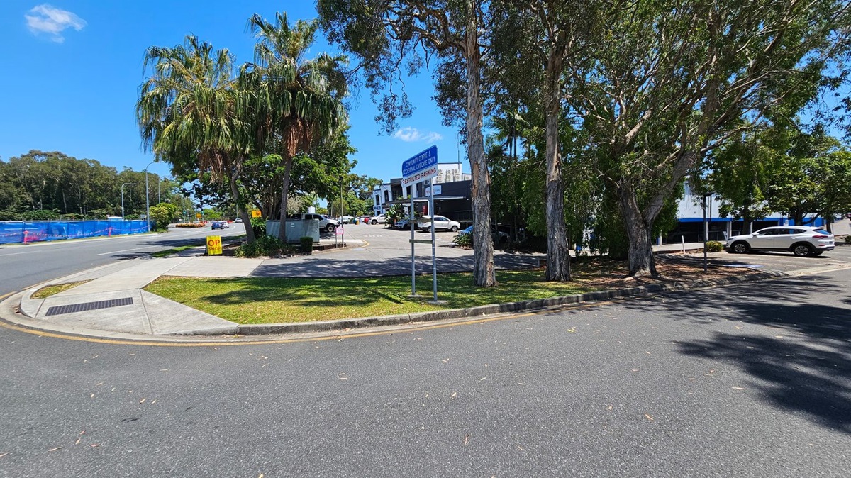 A street scene with a curved road in the foreground. On the left, there's a footpath and a grassy area with a sign that reads "OP SHOP." Palm trees and other greenery are present. In the centre, a parking area with several cars is visible, along with a blue sign indicating restricted parking. On the right, more trees provide shade, and a white car is parked. A building with a sign for a community centre and medical centre is in the background. A street scene with a curved road in the foreground. On the left, there's a footpath and a grassy area with a sign that reads "OP SHOP." Palm trees and other greenery are present. In the centre, a parking area with several cars is visible, along with a blue sign indicating restricted parking. On the right, more trees provide shade, and a white car is parked. A building with a sign for a community centre and medical centre is in the background.