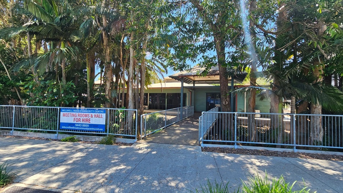 A community centre is partially obscured by tall trees and lush greenery. In the foreground, a concrete path leads to a ramp with metal railings, guiding towards the building's entrance. A sign on the left reads "Millwell Road Community Centre Meeting Rooms & Hall for Hire" with contact details. The building has a light green exterior with a covered entrance area. Shadows from the trees are cast on the path. A community centre is partially obscured by tall trees and lush greenery. In the foreground, a concrete path leads to a ramp with metal railings, guiding towards the building's entrance. A sign on the left reads "Millwell Road Community Centre Meeting Rooms & Hall for Hire" with contact details. The building has a light green exterior with a covered entrance area. Shadows from the trees are cast on the path.