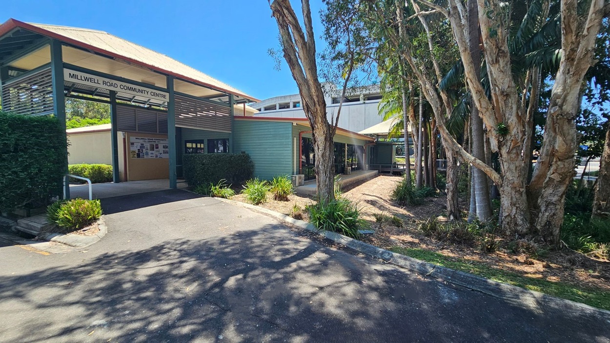 Millwell Road Community Centre with a sloped roof and a sign above the entrance. The building is light blue with a covered walkway. On the right, there are tall trees and a garden area with shrubs. The foreground shows a paved path with shadows from the trees. Millwell Road Community Centre with a sloped roof and a sign above the entrance. The building is light blue with a covered walkway. On the right, there are tall trees and a garden area with shrubs. The foreground shows a paved path with shadows from the trees.
