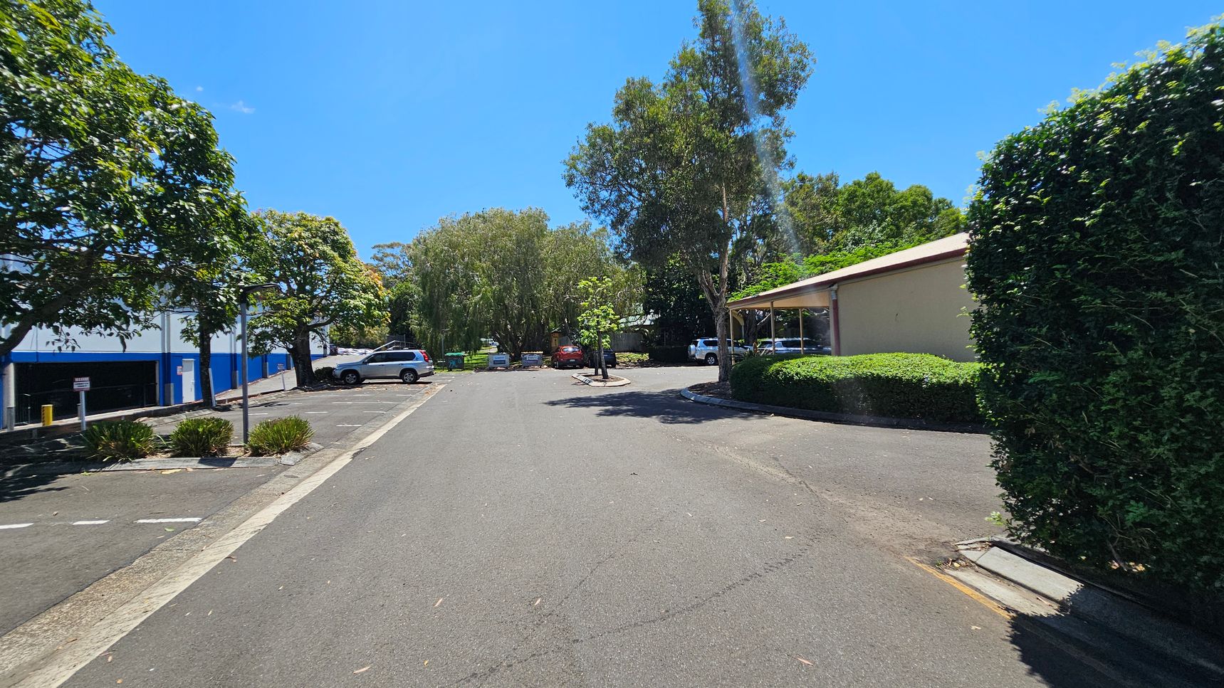 A car park with several vehicles is surrounded by trees and bushes. On the left, there is a blue and white building with a few parked cars. On the right, a beige building is partially visible, with a hedge and more parked cars. The ground is paved, and there are patches of grass and small plants along the edges. In the background, dense trees provide shade. A car park with several vehicles is surrounded by trees and bushes. On the left, there is a blue and white building with a few parked cars. On the right, a beige building is partially visible, with a hedge and more parked cars. The ground is paved, and there are patches of grass and small plants along the edges. In the background, dense trees provide shade.