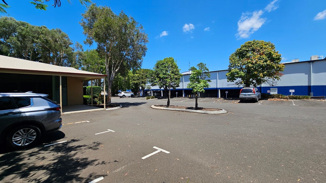 A car park with a few vehicles is visible. On the left, a grey car is parked near a building with a beige roof and green doors. In the centre, there are several trees planted in the parking area. On the right, a silver car is parked near a large building with a blue and white exterior. The ground is paved, and there are some leaves scattered around. Trees and greenery are in the background. A car park with a few vehicles is visible. On the left, a grey car is parked near a building with a beige roof and green doors. In the centre, there are several trees planted in the parking area. On the right, a silver car is parked near a large building with a blue and white exterior. The ground is paved, and there are some leaves scattered around. Trees and greenery are in the background.