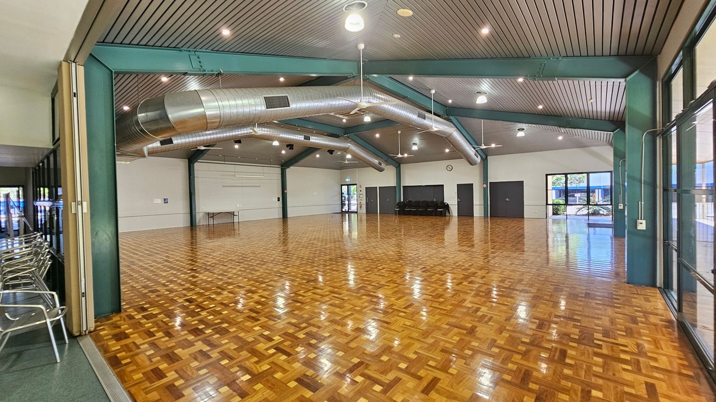 A spacious hall with a polished parquet floor and a high ceiling featuring exposed metal beams and large air ducts. On the left, there are stacked metal chairs against a wall. The back wall has a clock and several doors, with a small table nearby. Large windows on the right allow natural light to enter, showing a glimpse of greenery outside. The ceiling has multiple lights and fans. A spacious hall with a polished parquet floor and a high ceiling featuring exposed metal beams and large air ducts. On the left, there are stacked metal chairs against a wall. The back wall has a clock and several doors, with a small table nearby. Large windows on the right allow natural light to enter, showing a glimpse of greenery outside. The ceiling has multiple lights and fans.