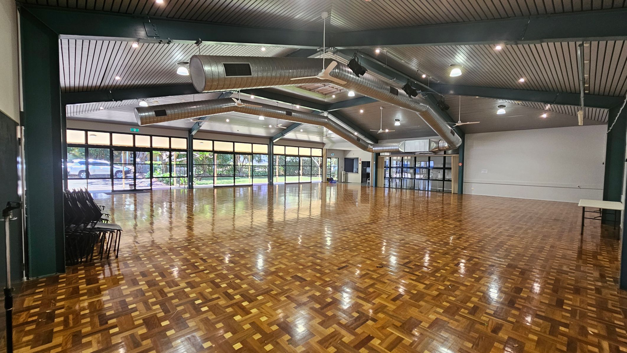 A spacious hall with a polished parquet floor. On the left, there are stacked chairs against the wall. The right side features a plain white wall with a small table. The background has large windows and glass doors, allowing natural light to enter and providing a view of greenery and parked cars outside. The ceiling is lined with exposed ductwork and lighting fixtures. A spacious hall with a polished parquet floor. On the left, there are stacked chairs against the wall. The right side features a plain white wall with a small table. The background has large windows and glass doors, allowing natural light to enter and providing a view of greenery and parked cars outside. The ceiling is lined with exposed ductwork and lighting fixtures.
