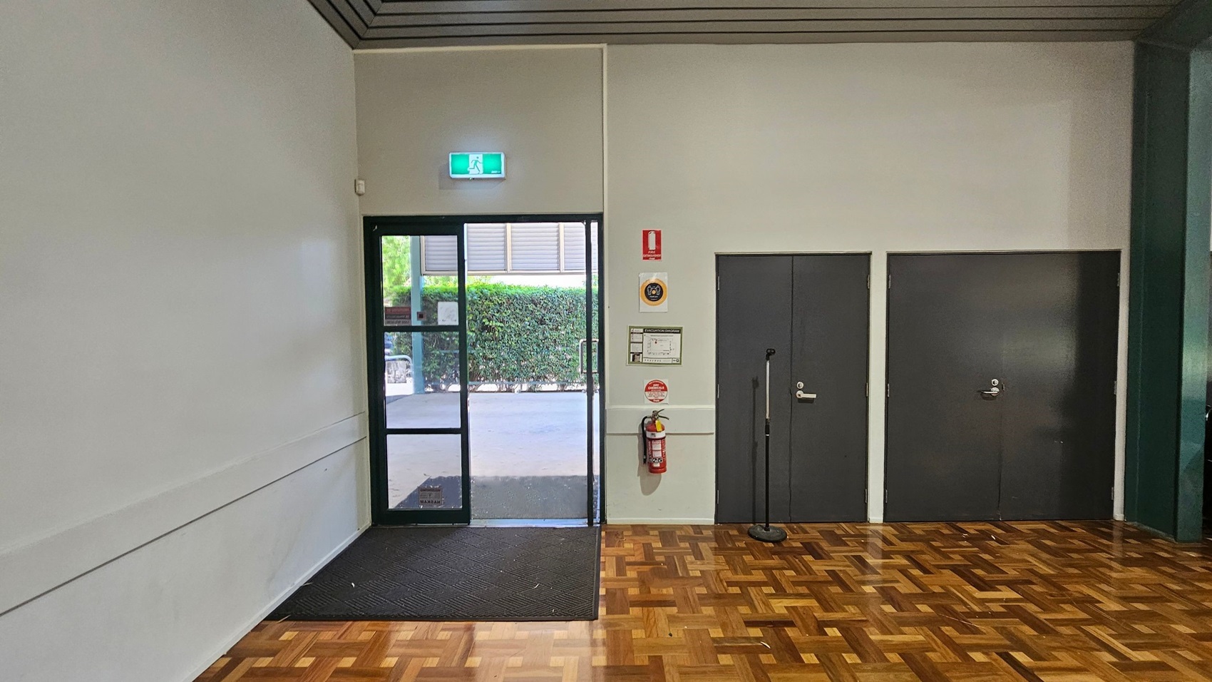 A room with a parquet wooden floor and a light green ceiling. On the left, there's a glass door with an illuminated exit sign above it, leading outside where greenery is visible. To the right of the door, there are two dark grey doors. A fire extinguisher is mounted on the wall between the glass door and the grey doors, along with some safety signs. A black mat is placed in front of the glass door. A room with a parquet wooden floor and a light green ceiling. On the left, there's a glass door with an illuminated exit sign above it, leading outside where greenery is visible. To the right of the door, there are two dark grey doors. A fire extinguisher is mounted on the wall between the glass door and the grey doors, along with some safety signs. A black mat is placed in front of the glass door.