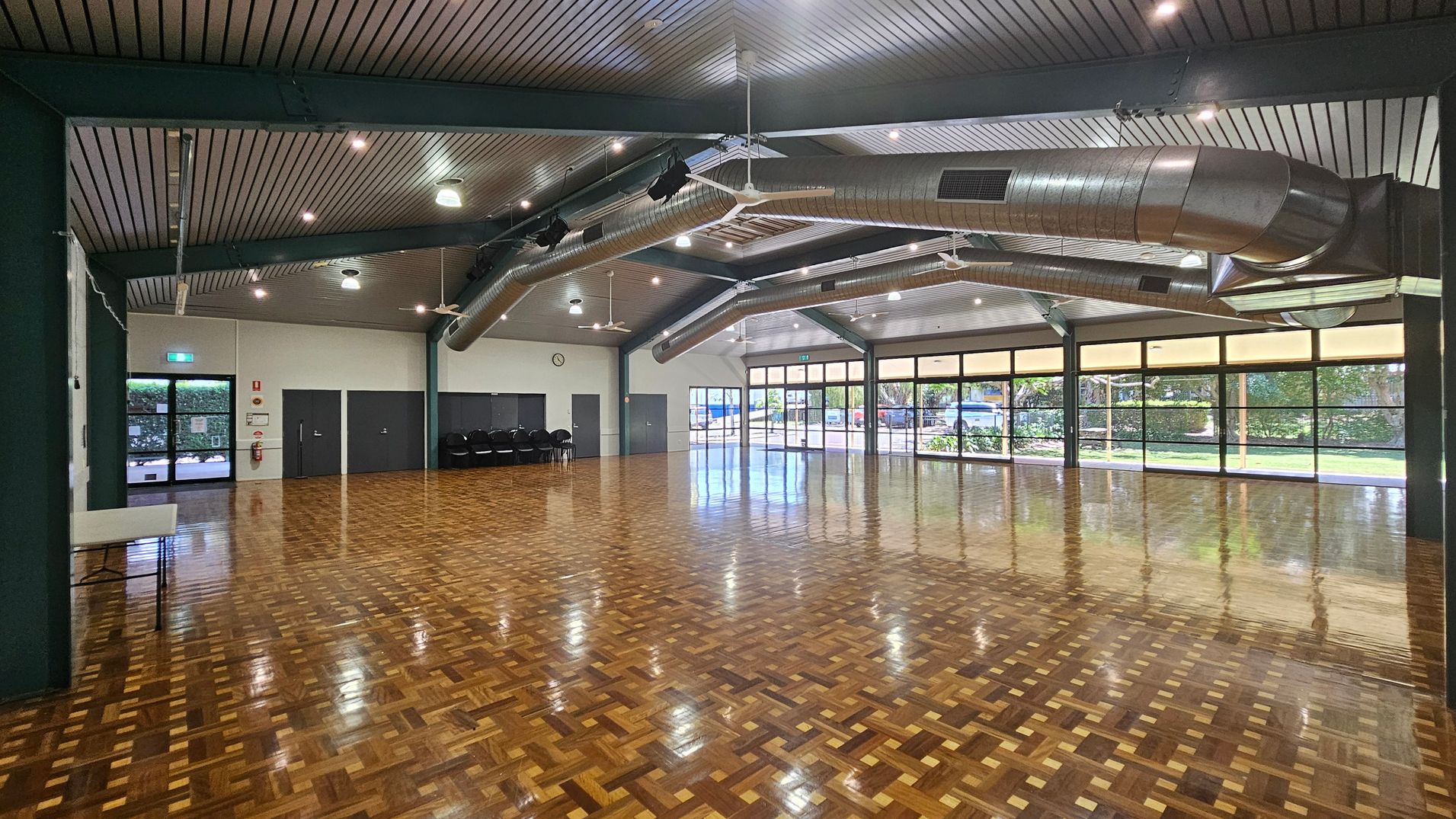 A spacious hall with a polished parquet floor and a high, slatted ceiling featuring exposed ductwork and ceiling fans. On the left, there are dark double doors and a stack of chairs against the wall. The right side has large windows with views of greenery outside. The ceiling has multiple lights, and the walls are light-coloured. A clock is mounted on the wall near the doors. A spacious hall with a polished parquet floor and a high, slatted ceiling featuring exposed ductwork and ceiling fans. On the left, there are dark double doors and a stack of chairs against the wall. The right side has large windows with views of greenery outside. The ceiling has multiple lights, and the walls are light-coloured. A clock is mounted on the wall near the doors.