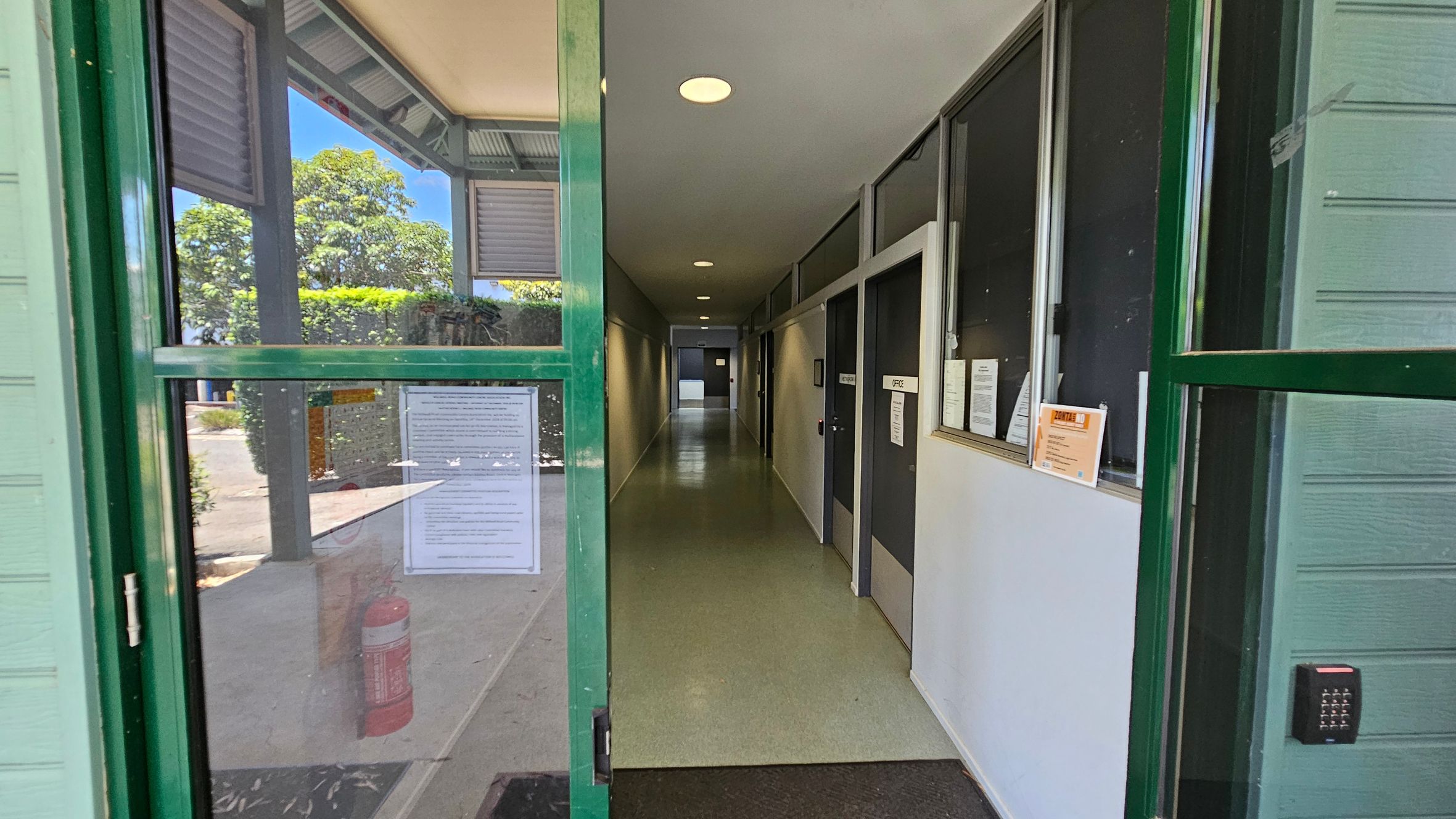 A set of green-framed glass doors, one open, leads into a building with light green wooden panelling. The floor outside is covered with a dark mat and scattered leaves. To the left, a beige wall displays a poster. Inside, a corridor with a green floor is visible. On the right, a small bush is next to a railing. A plaque is mounted on the wall above the bush. A set of green-framed glass doors, one open, leads into a building with light green wooden panelling. The floor outside is covered with a dark mat and scattered leaves. To the left, a beige wall displays a poster. Inside, a corridor with a green floor is visible. On the right, a small bush is next to a railing. A plaque is mounted on the wall above the bush.