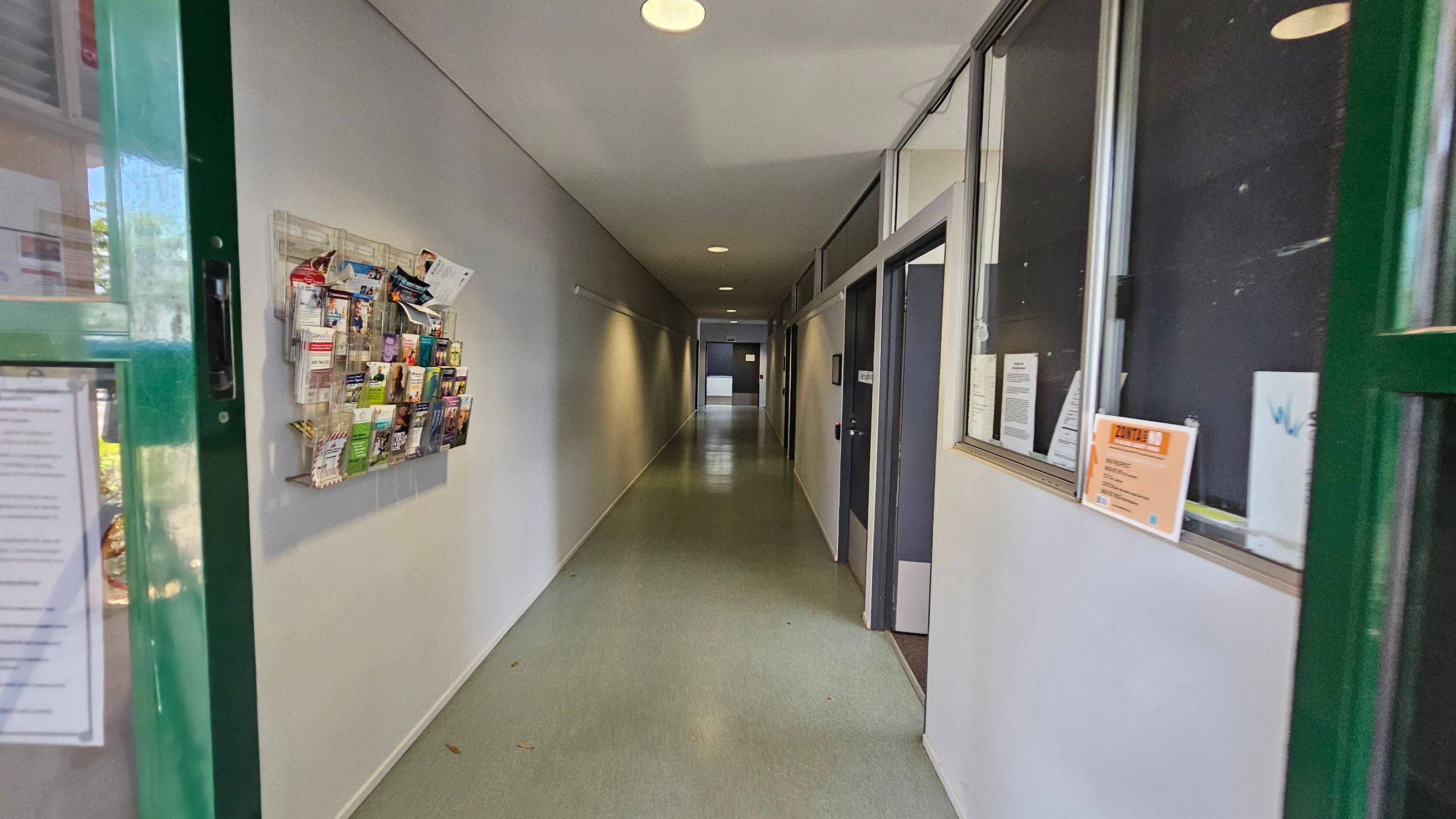 A hallway with a green door partially open on the left. A wall-mounted rack filled with brochures is on the left wall. The floor is light green, and the ceiling has recessed lights. On the right, there are windows with notices and a door. The corridor extends into the distance, with more doors and a light at the end. A hallway with a green door partially open on the left. A wall-mounted rack filled with brochures is on the left wall. The floor is light green, and the ceiling has recessed lights. On the right, there are windows with notices and a door. The corridor extends into the distance, with more doors and a light at the end.