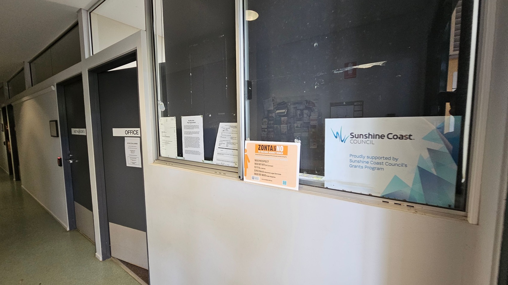 A corridor with a green floor and light-coloured walls. On the left, there are two dark doors labelled "OFFICE" and "ENTRY/EXIT". On the right, a window displays several notices, including a "Zonta Says No" poster and a "Sunshine Coast Council" sign, indicating support from the council's grants programme. The ceiling is not visible. A corridor with a green floor and light-coloured walls. On the left, there are two dark doors labelled "OFFICE" and "ENTRY/EXIT". On the right, a window displays several notices, including a "Zonta Says No" poster and a "Sunshine Coast Council" sign, indicating support from the council's grants programme. The ceiling is not visible.