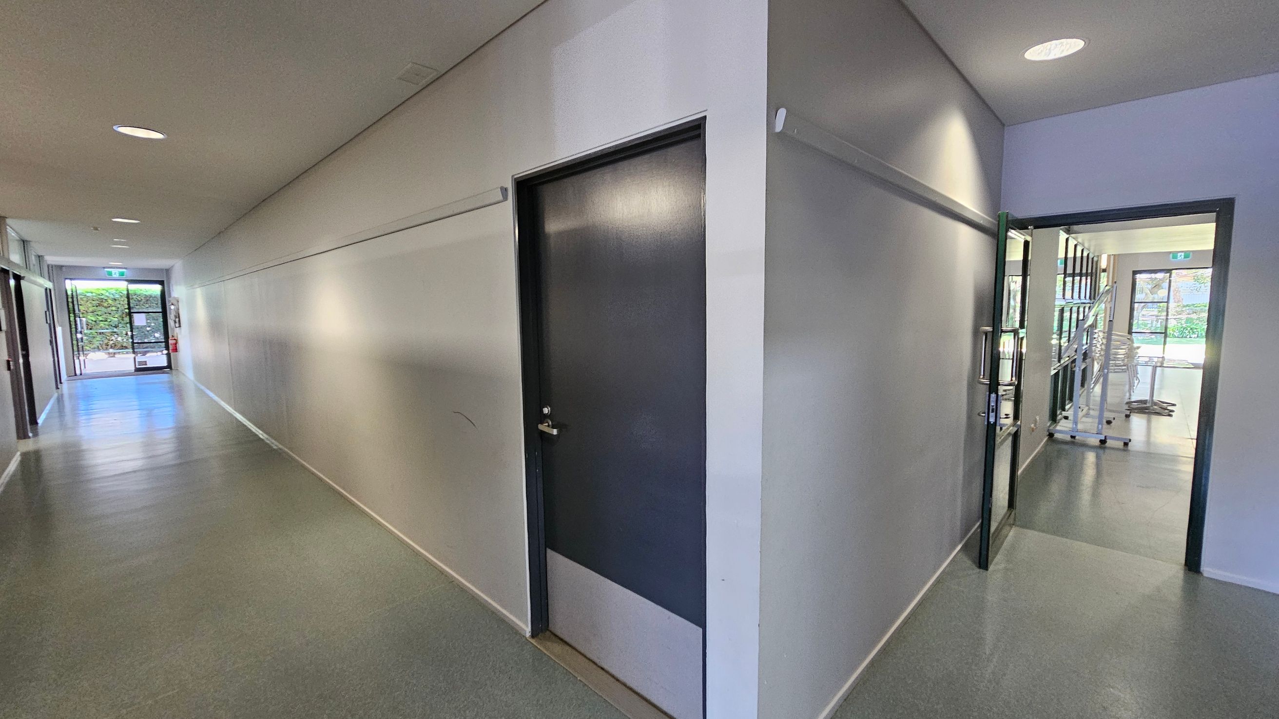 A corridor with grey flooring and white walls. On the left, a closed dark door is set into the wall. The corridor extends towards a glass door at the end, leading outside with greenery visible. On the right, an open door leads into a room with shelves and stacked chairs. The ceiling has recessed lighting. A corridor with grey flooring and white walls. On the left, a closed dark door is set into the wall. The corridor extends towards a glass door at the end, leading outside with greenery visible. On the right, an open door leads into a room with shelves and stacked chairs. The ceiling has recessed lighting.