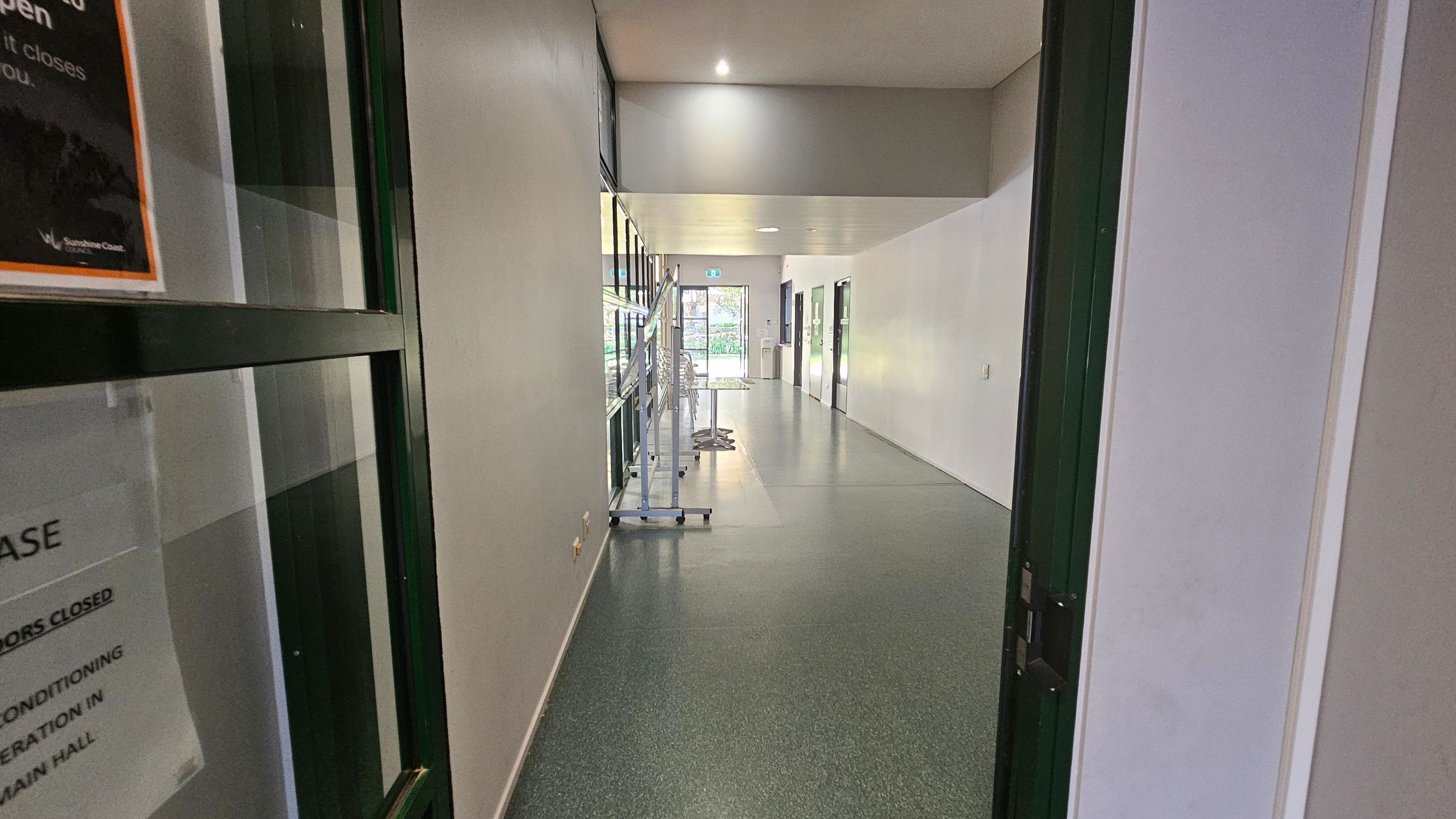 A hallway with a speckled grey floor and white walls. On the left, there's a glass door with a sign. Further down the left side, a row of windows and a metal rack with stacked chairs. On the right, several closed doors line the wall. At the end of the hallway, a glass door leads outside, with visible greenery through it. The ceiling is plain with recessed lighting. A hallway with a speckled grey floor and white walls. On the left, there's a glass door with a sign. Further down the left side, a row of windows and a metal rack with stacked chairs. On the right, several closed doors line the wall. At the end of the hallway, a glass door leads outside, with visible greenery through it. The ceiling is plain with recessed lighting.