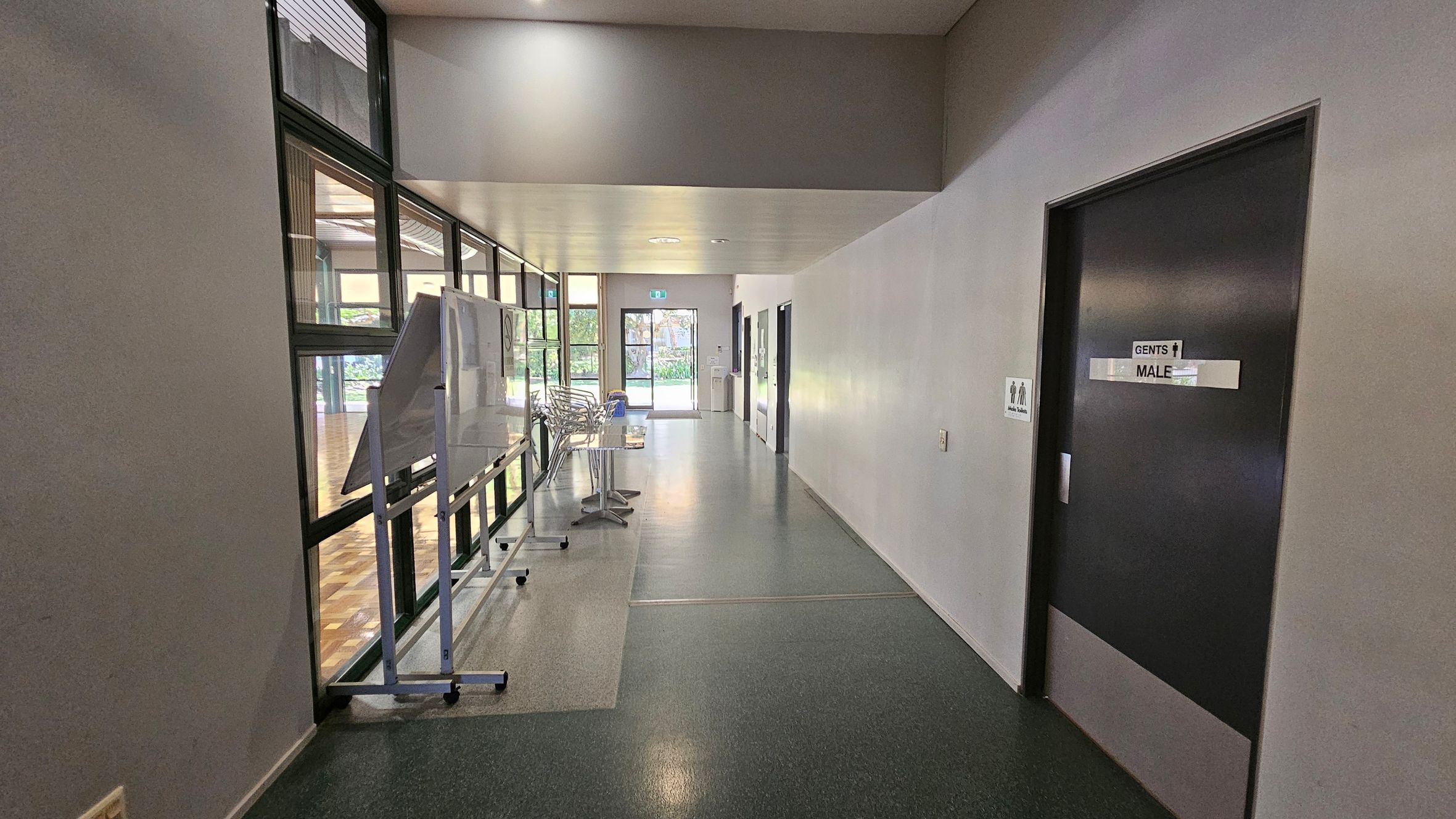 A hallway with a dark floor and light ceiling. On the right, there's a door marked "Gents Male" with a sign next to it. On the left, large windows reveal a wooden-floored room. A whiteboard and stacked chairs are positioned along the windows. At the end of the hallway, a glass door leads outside, with greenery visible through it. A hallway with a dark floor and light ceiling. On the right, there's a door marked "Gents Male" with a sign next to it. On the left, large windows reveal a wooden-floored room. A whiteboard and stacked chairs are positioned along the windows. At the end of the hallway, a glass door leads outside, with greenery visible through it.