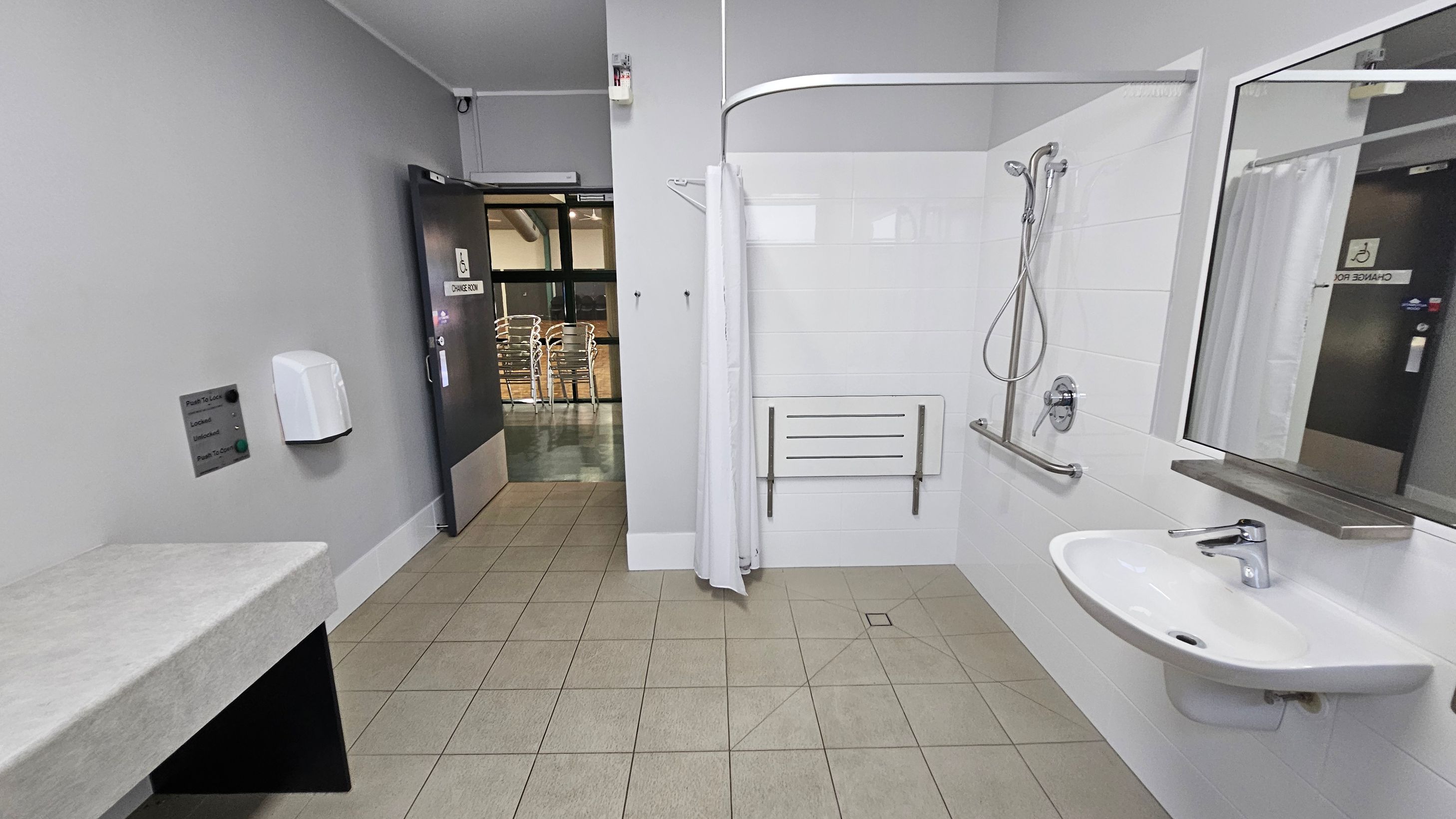 A spacious, accessible bathroom with a light green tiled floor. On the right, there's a white sink with a mirror above it and a shower area with a handheld showerhead, grab bar, and a foldable seat. A white curtain partially encloses the shower. On the left, a grey wall features a soap dispenser and a control panel. The door is open, revealing a hallway with stacked chairs visible outside. The ceiling is plain and white. A spacious, accessible bathroom with a light green tiled floor. On the right, there's a white sink with a mirror above it and a shower area with a handheld showerhead, grab bar, and a foldable seat. A white curtain partially encloses the shower. On the left, a grey wall features a soap dispenser and a control panel. The door is open, revealing a hallway with stacked chairs visible outside. The ceiling is plain and white.