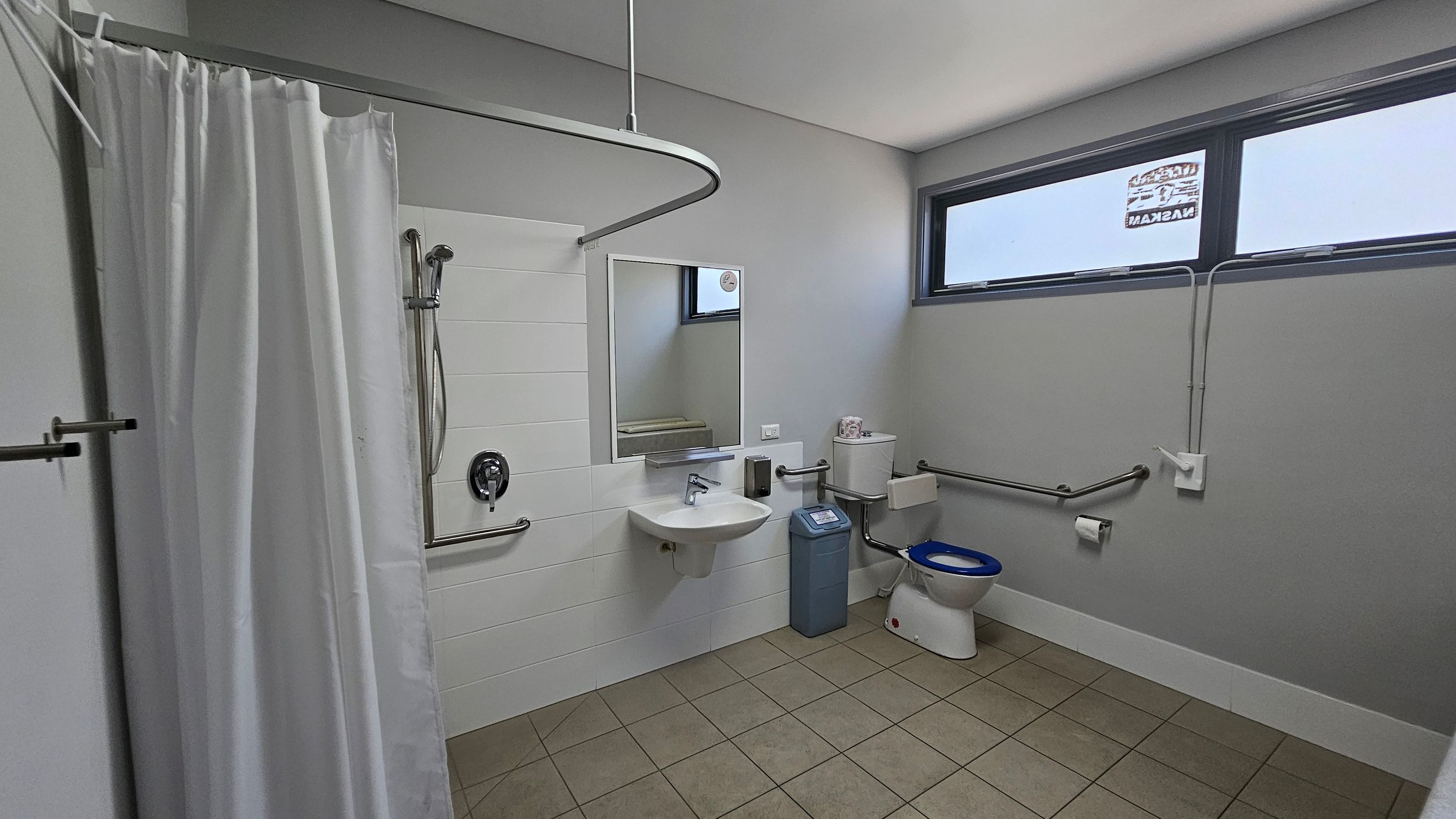 A bathroom with a green tiled floor and a white ceiling. On the left, there's a shower area with a white curtain and a wall-mounted showerhead. In the centre, a wall-mounted sink with a mirror above it is flanked by a soap dispenser. To the right, a toilet with a blue seat is next to a blue bin. The room has grab bars for accessibility and a high horizontal window on the right wall. A bathroom with a green tiled floor and a white ceiling. On the left, there's a shower area with a white curtain and a wall-mounted showerhead. In the centre, a wall-mounted sink with a mirror above it is flanked by a soap dispenser. To the right, a toilet with a blue seat is next to a blue bin. The room has grab bars for accessibility and a high horizontal window on the right wall.