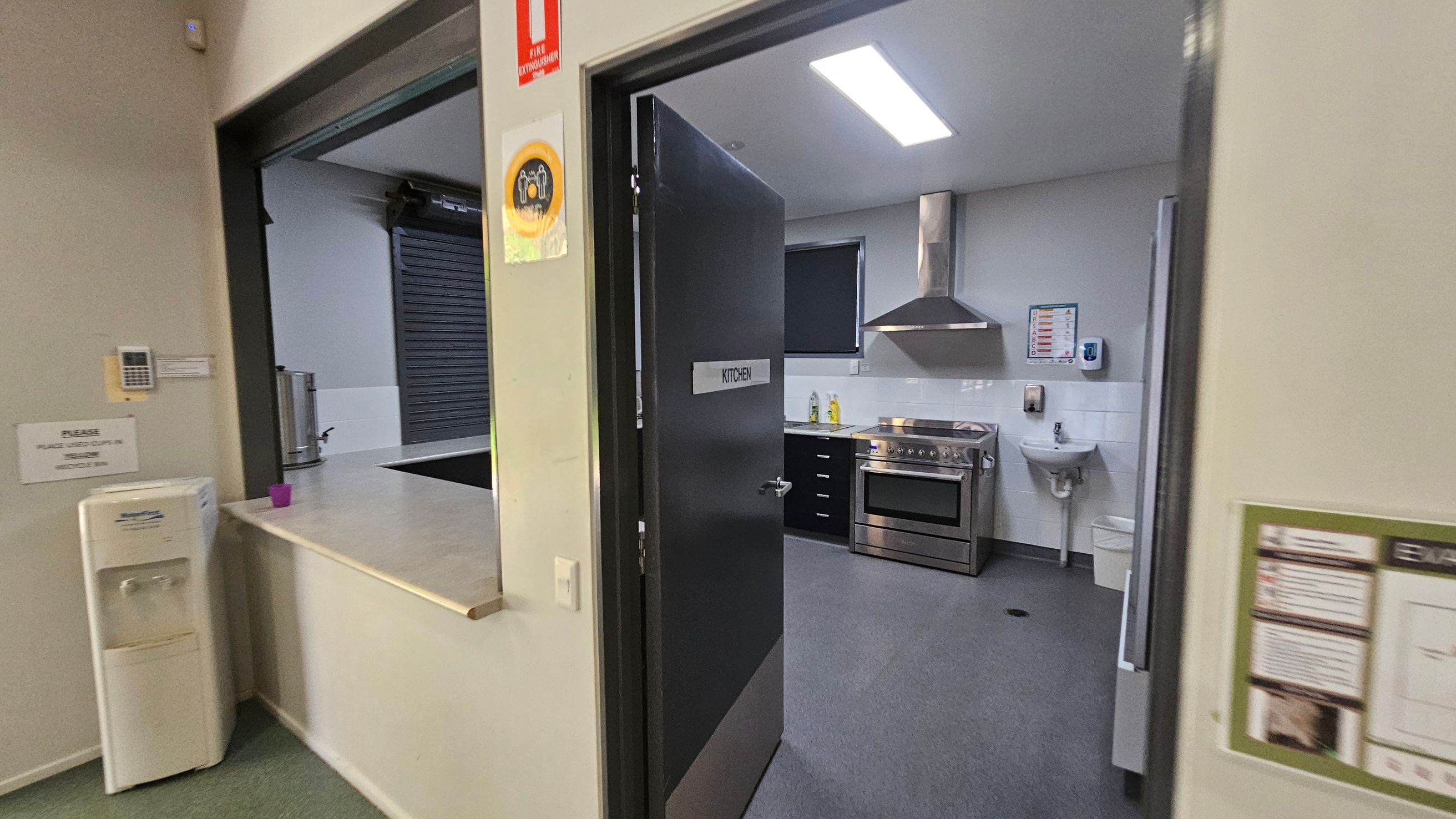 A kitchen with a black door labelled "KITCHEN" is open, revealing a stainless steel oven and range hood. A small sink is mounted on the wall to the right. On the left, a counter with a water cooler and a purple cup is visible. A fire extinguisher sign is above the door. The floor is grey, and the ceiling has a bright fluorescent light. A kitchen with a black door labelled "KITCHEN" is open, revealing a stainless steel oven and range hood. A small sink is mounted on the wall to the right. On the left, a counter with a water cooler and a purple cup is visible. A fire extinguisher sign is above the door. The floor is grey, and the ceiling has a bright fluorescent light.
