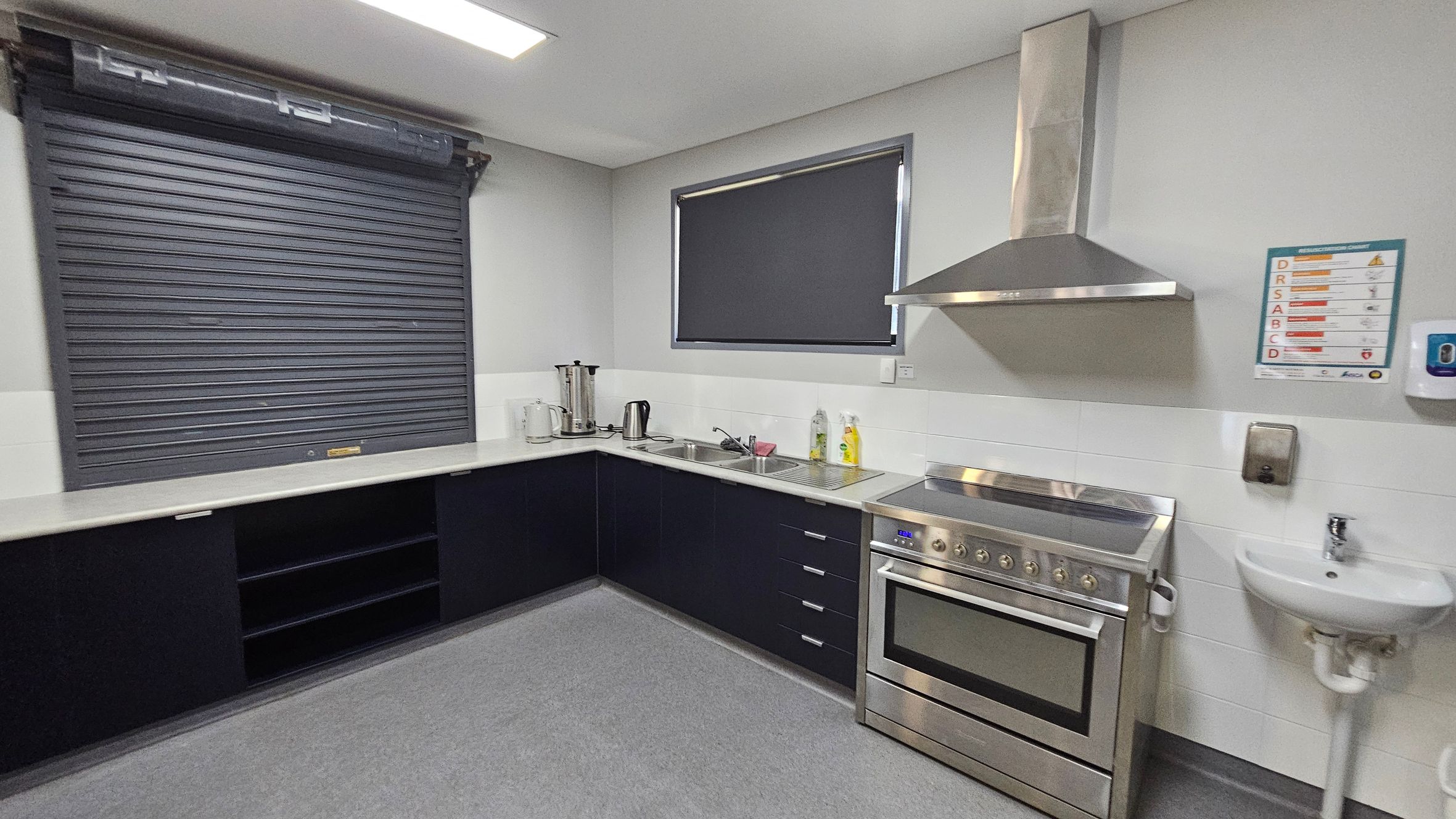 A modern kitchen with a grey roller shutter on the left above black cabinets. In the middle, there's a countertop with a kettle, a coffee maker, and a sink. On the right, a stainless steel oven with an overhead extractor fan is next to a small white sink. A window with a dark blind is above the counter. The floor is grey, and the ceiling has a rectangular light fixture. A safety instruction poster and a soap dispenser are mounted on the wall. A modern kitchen with a grey roller shutter on the left above black cabinets. In the middle, there's a countertop with a kettle, a coffee maker, and a sink. On the right, a stainless steel oven with an overhead extractor fan is next to a small white sink. A window with a dark blind is above the counter. The floor is grey, and the ceiling has a rectangular light fixture. A safety instruction poster and a soap dispenser are mounted on the wall.